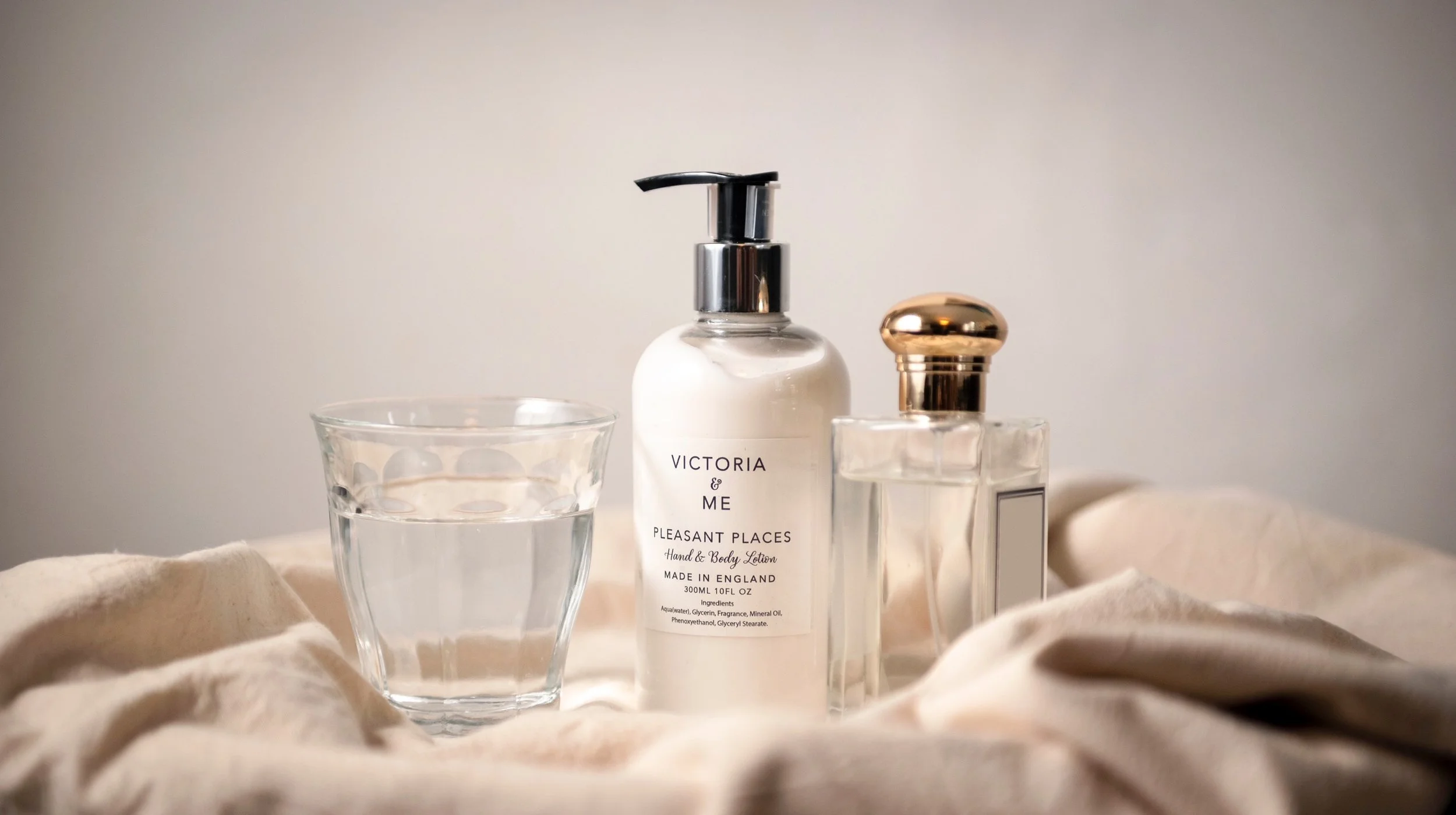 A clear glass of water, a white lotion or soap bottle with a black pump labeled 'Victoria & Me Pleasant Places Hand & Body Lotion', and a transparent perfume or cosmetic bottle with a gold cap are arranged on a soft, beige cloth.