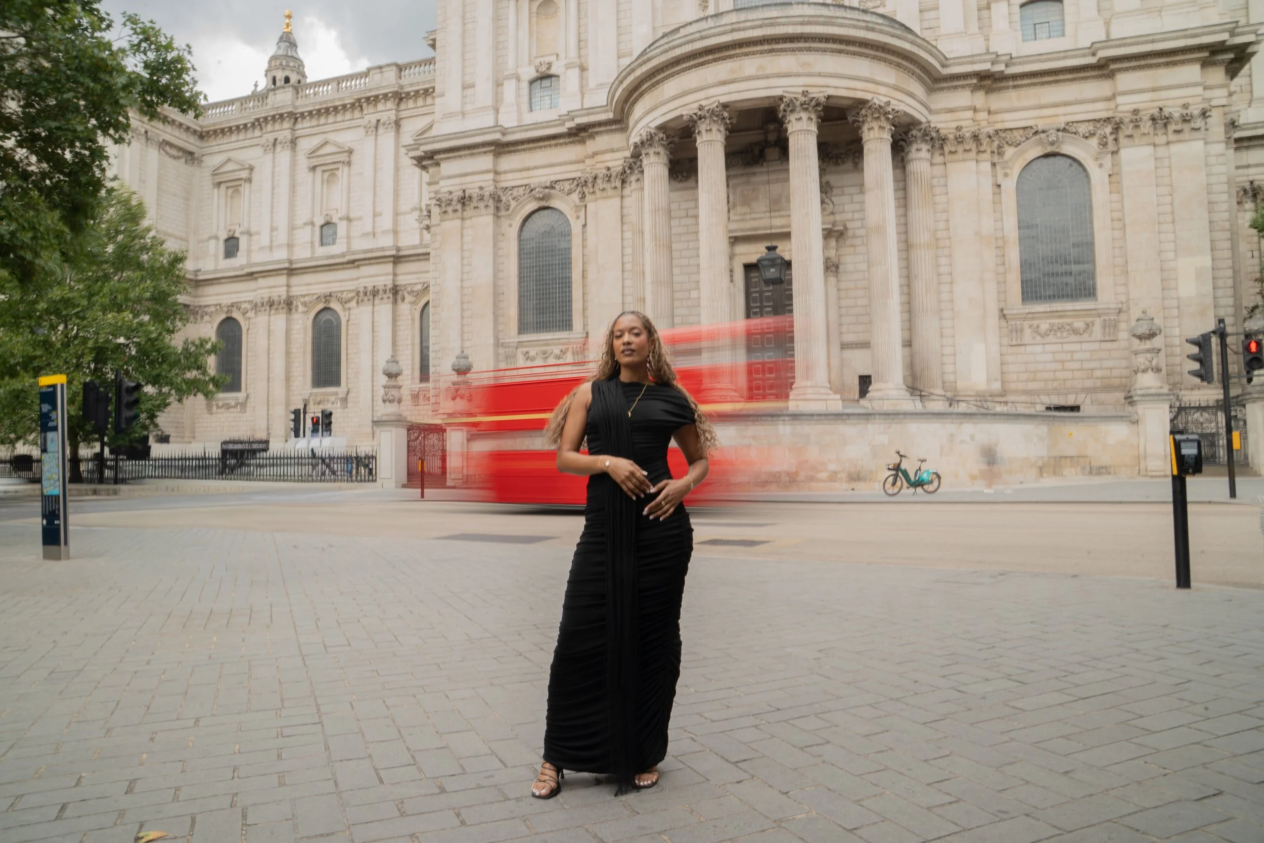 A woman in a black dress and jewellery from Onna London standing in front of a historic building with classical architecture in an urban setting in Central London. A red double-decker bus appears as a motion blur in the background.