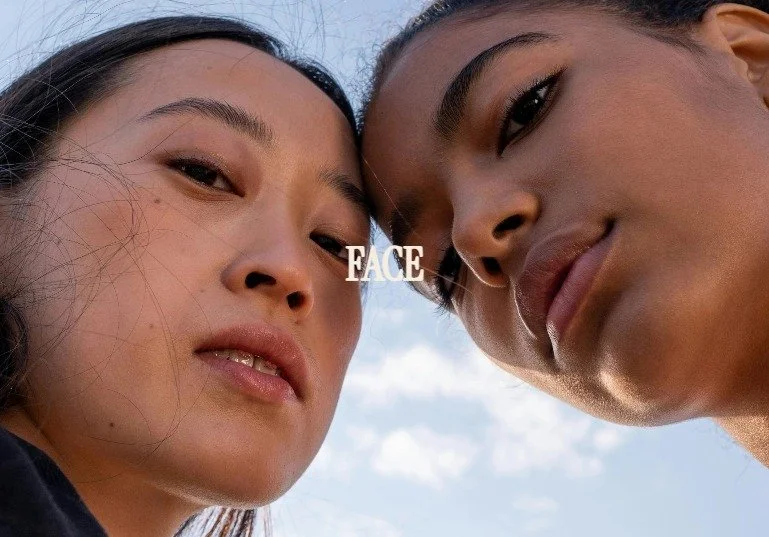 Close-up of two young women looking down at the camera, with the word 'FACE' between them, against a partly cloudy sky.