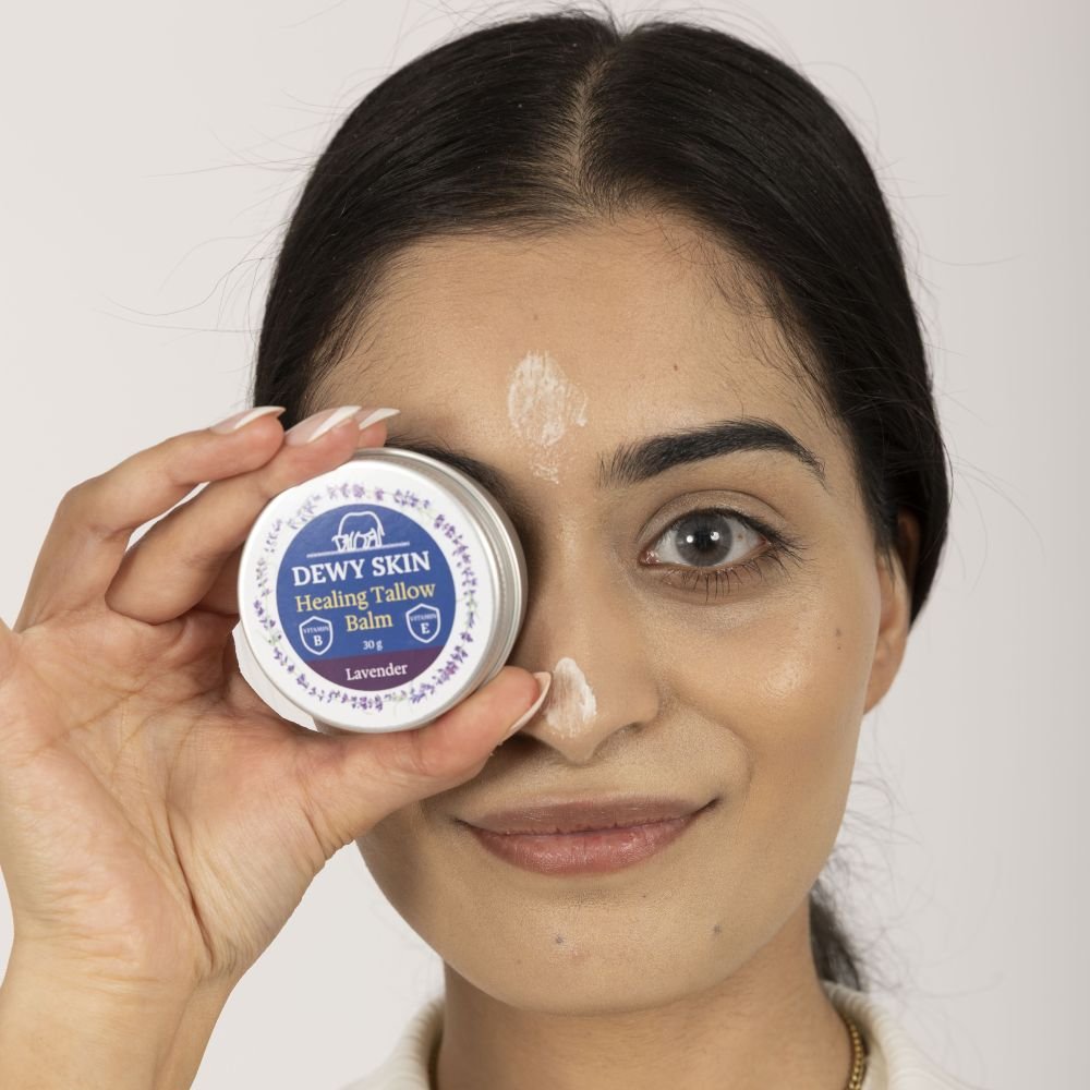 A woman with dark hair and light skin holds a small tin of Dewy Skin Healing Tallow Balm with lavender up to her face, covering her right eye, and smiling. She has white cream on her forehead and nose.