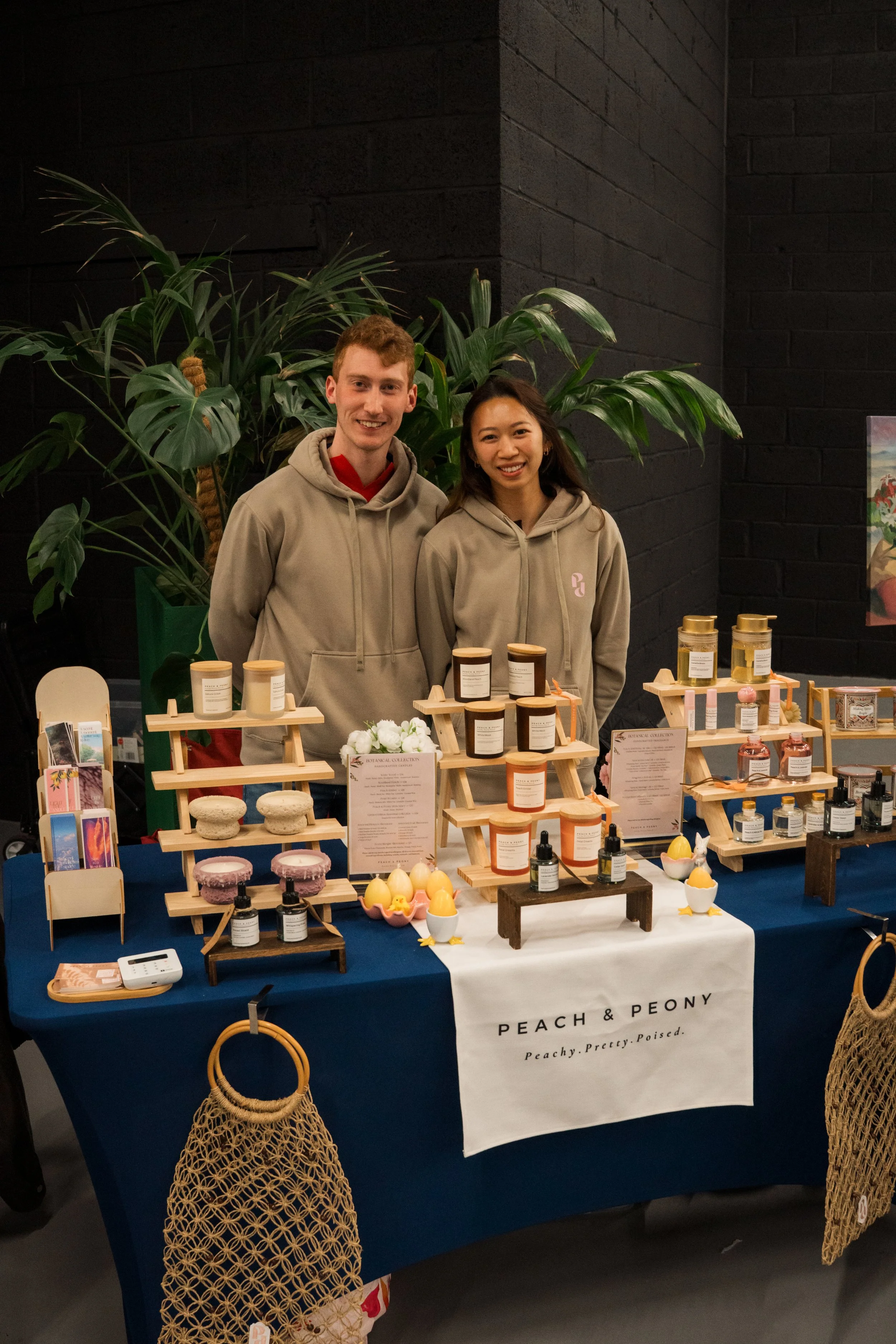 Two people standing behind a booth displaying skincare products, with a large green plant behind them, and items like soaps, lotions, and small jars on the table.