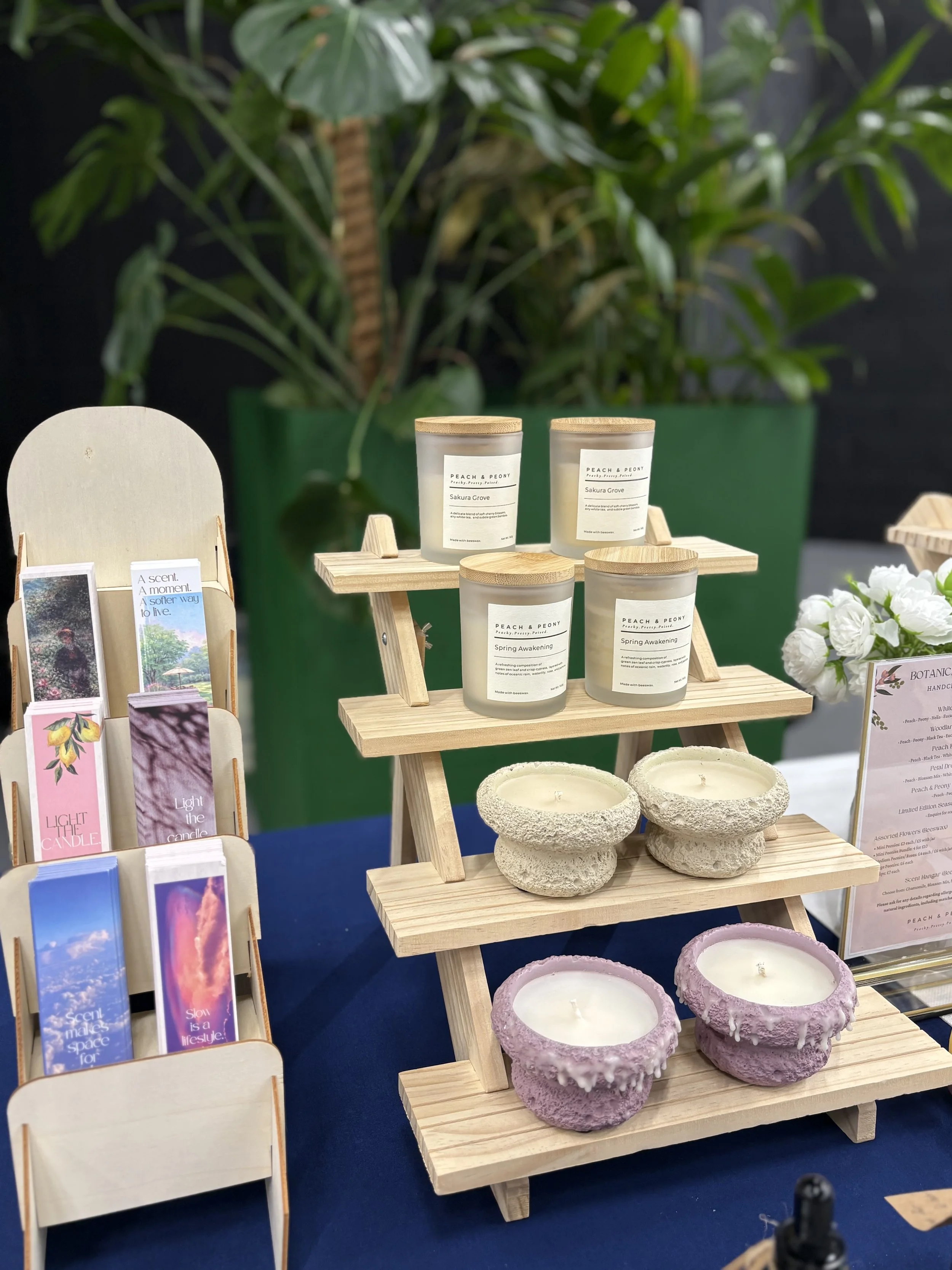 Display of scented candles, wooden trays, and decorative plant pots on a woodenstand, with a backdrop of green plants.