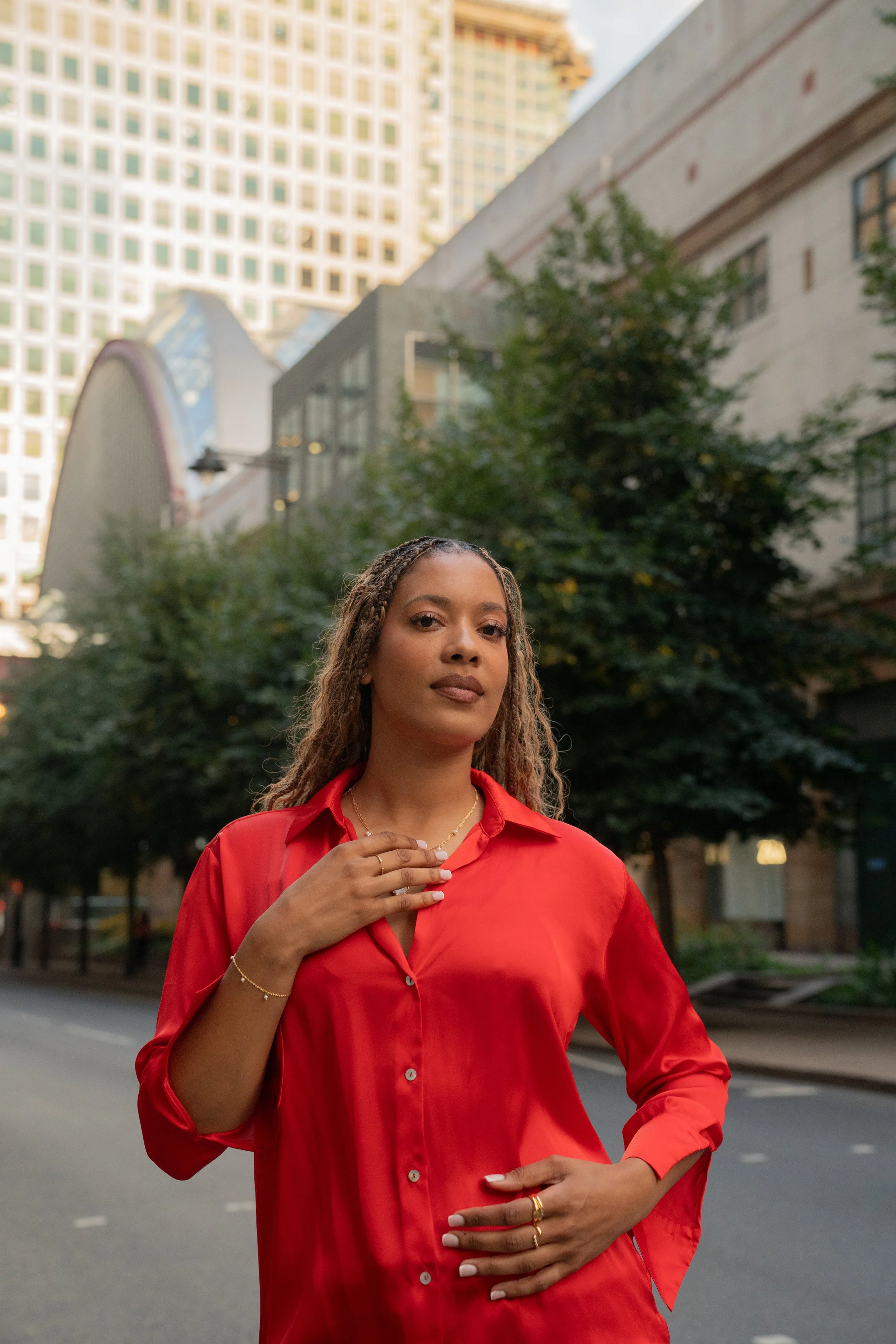 A woman in a red shirt and jewellery from Onna London standing outdoors in an urban area within London with tall buildings and trees in the background.