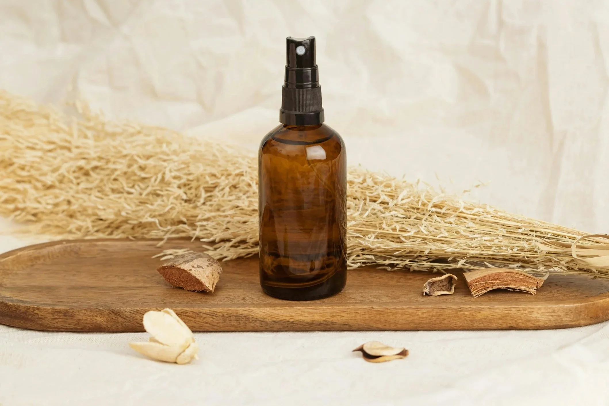 A brown glass spray bottle on a wooden tray with dried herbs and plant pieces surrounding it, with a background of straw-like material.