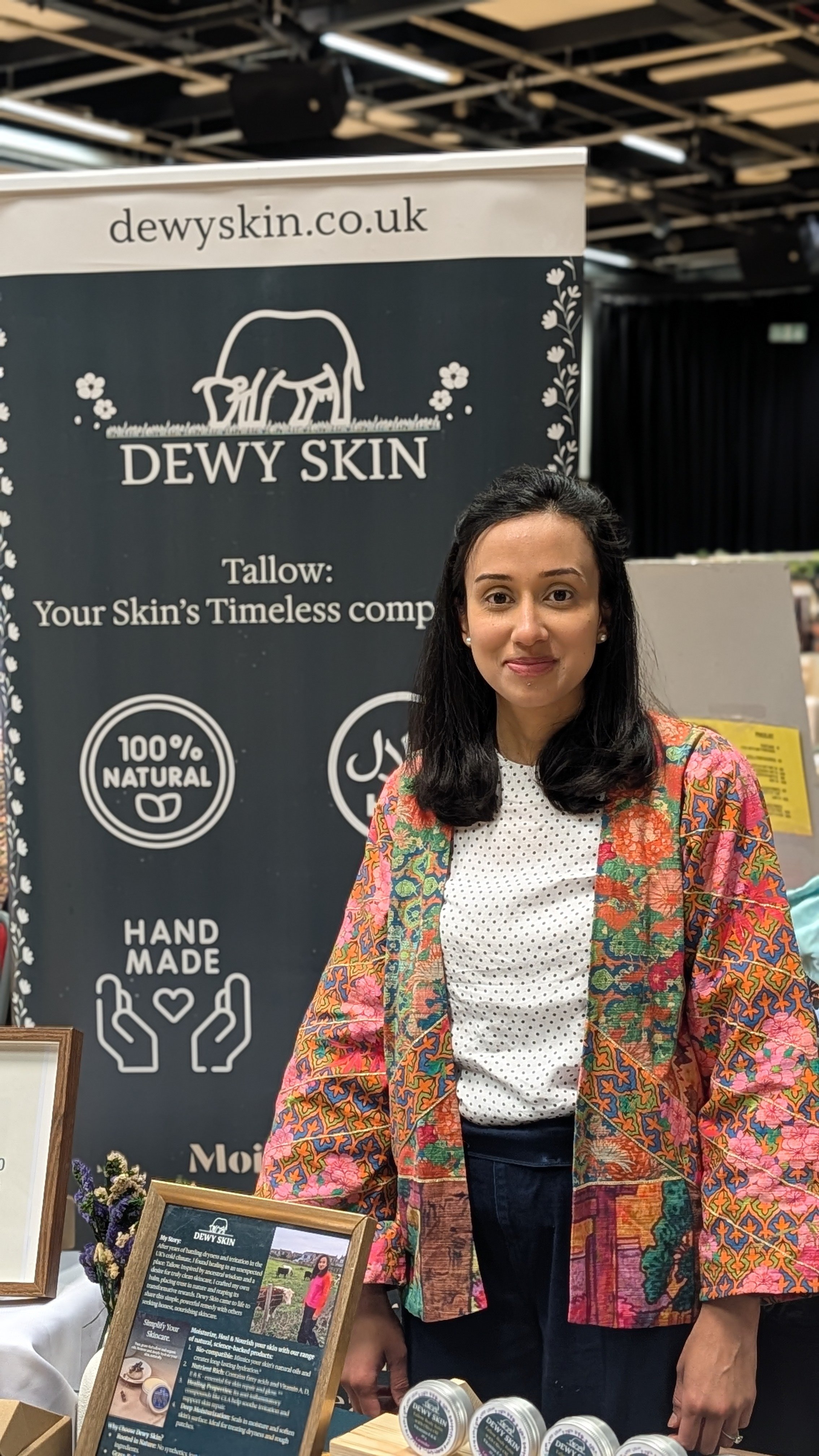 Woman, founder of Dewy Skin, standing behind a booth at an event, wearing a colorful patterned jacket, with a Dewy Skin banner in the background and skincare products on display.
