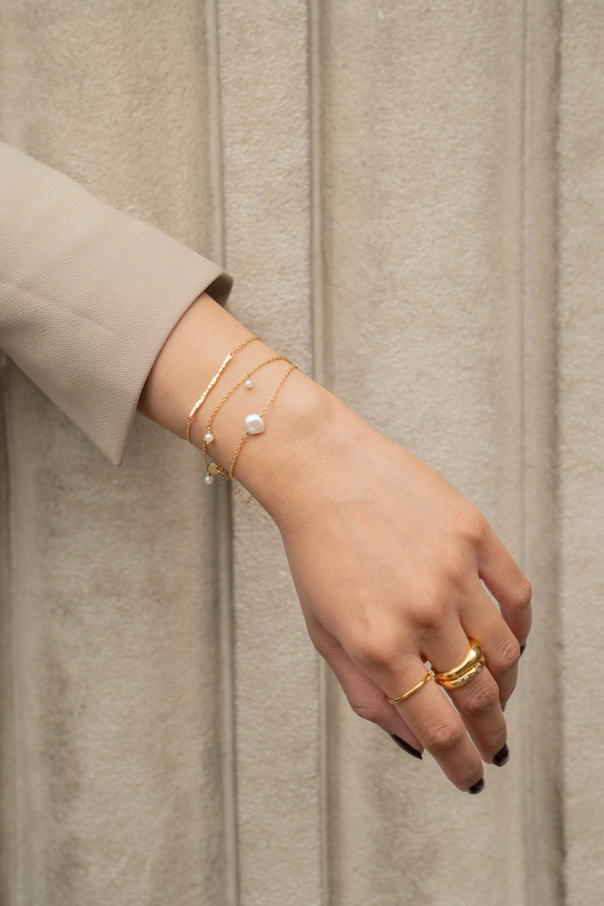 A person's hand includes multiple gold rings and delicate gold bracelets with pearls from Onna London, resting against a beige stone textured background.