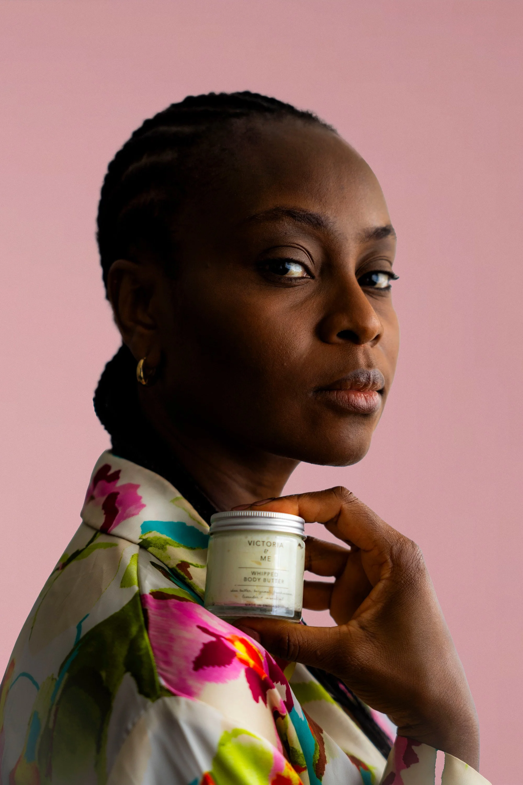 A woman with dark skin and braided hair holding a jar of whipped body butter, looking to the side against a pink background. She wears a colorful floral-patterned garment and gold earrings.