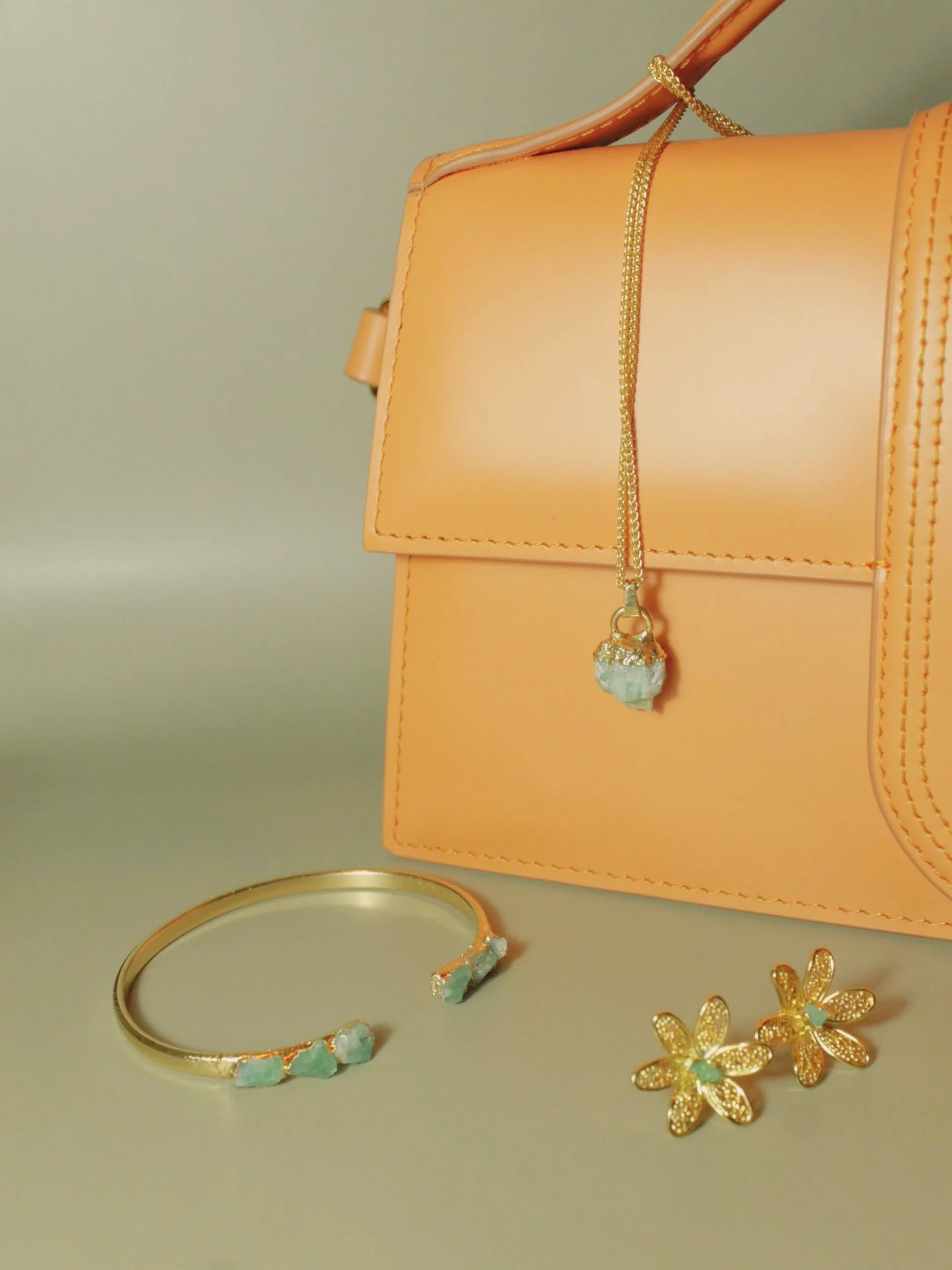 Gold jewelry including a necklace with a large pendant, earrings and a bracelet, displayed against a light-colored background and a handbag.