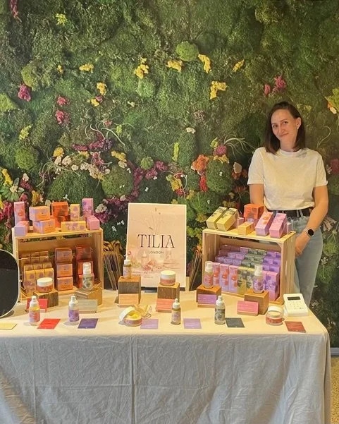 A woman, founder of Tilia London, standing behind a table displaying skincare or cosmetic products at a booth with a lush green floral mural background.