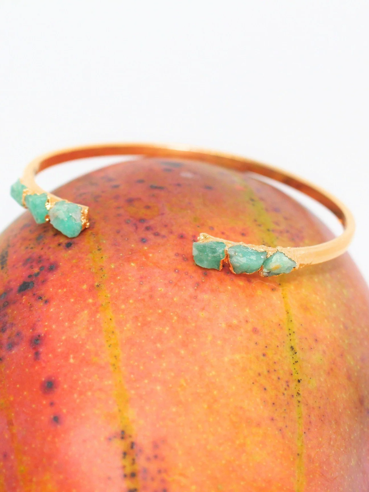 Gold bracelet with light green, rough gemstone, emerald, accents resting on a red and yellow speckled apple.