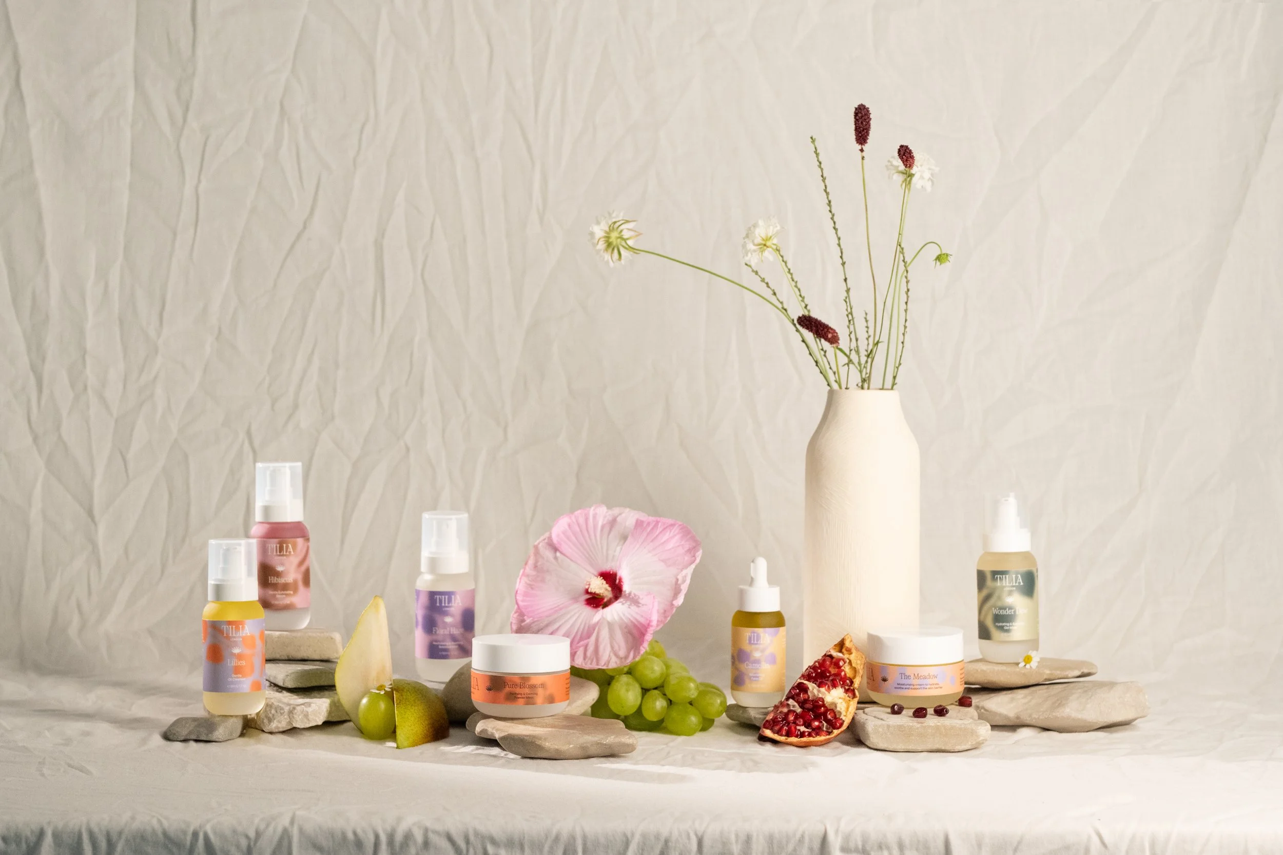 A flat lay of Tilia London skincare products with a vase of flowers and pomegranate on a textured white background.