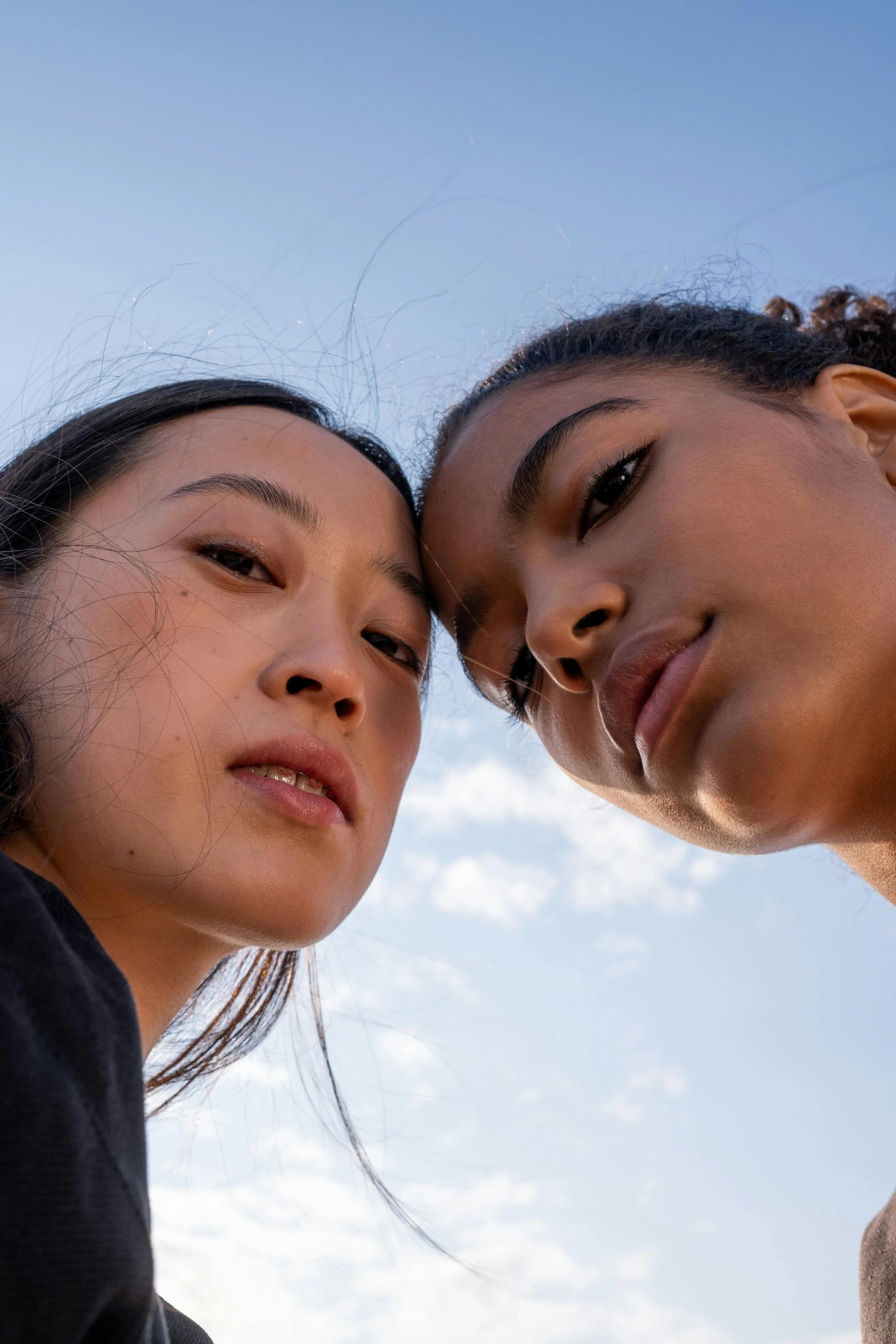 Close-up of two young women looking downward, their faces near each other, against a blue sky with some clouds.