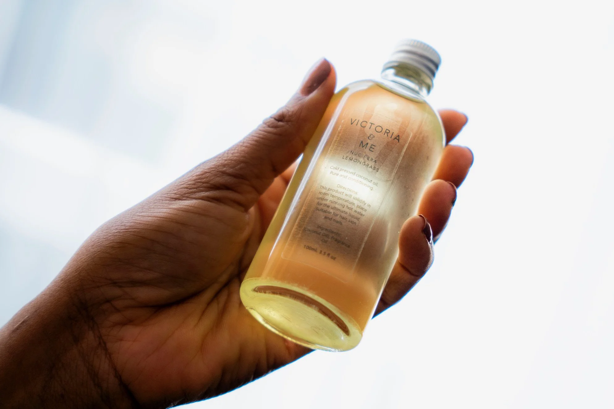 A hand holding a transparent bottle of coconut oil infused with lemon grass named Victoria & Me, with a white background.