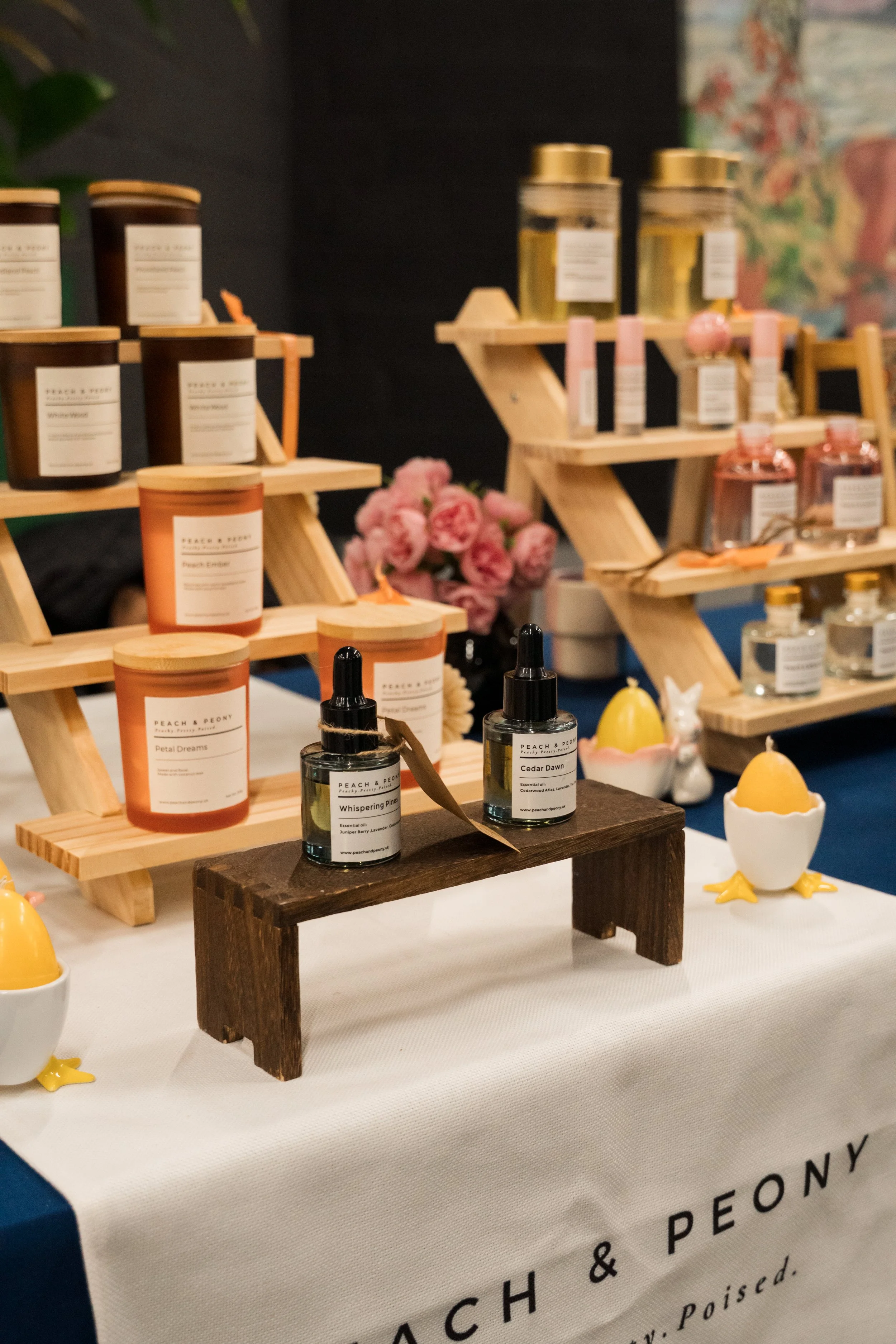 Display of scented candles, essential oils, and decorations on wooden shelves and a table, with pink flowers and small decorative items.