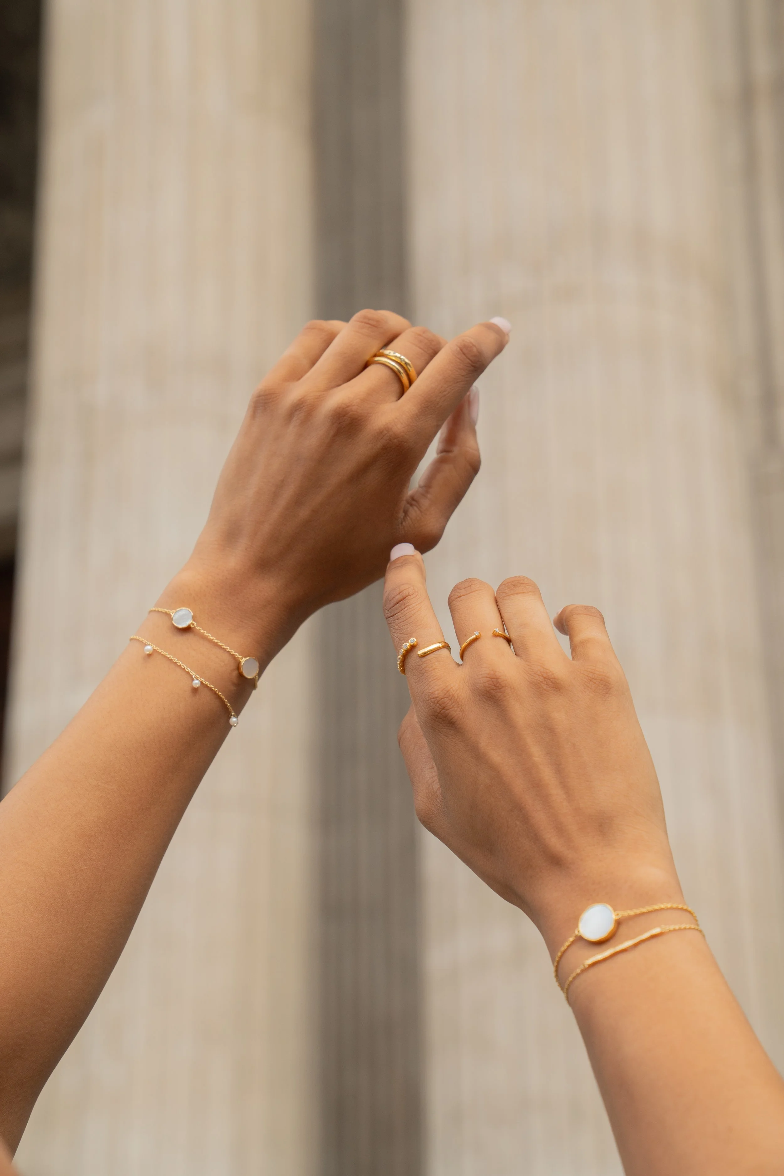 Two hands are shown with multiple jewelry pieces including rings and bracelets from Onna London, with a blurred background featuring roman style pillars in central London.