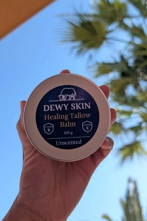 Hand holding a round container of Dewy Skin Healing Tallow Balm outdoors with palm trees and blue sky in the background.