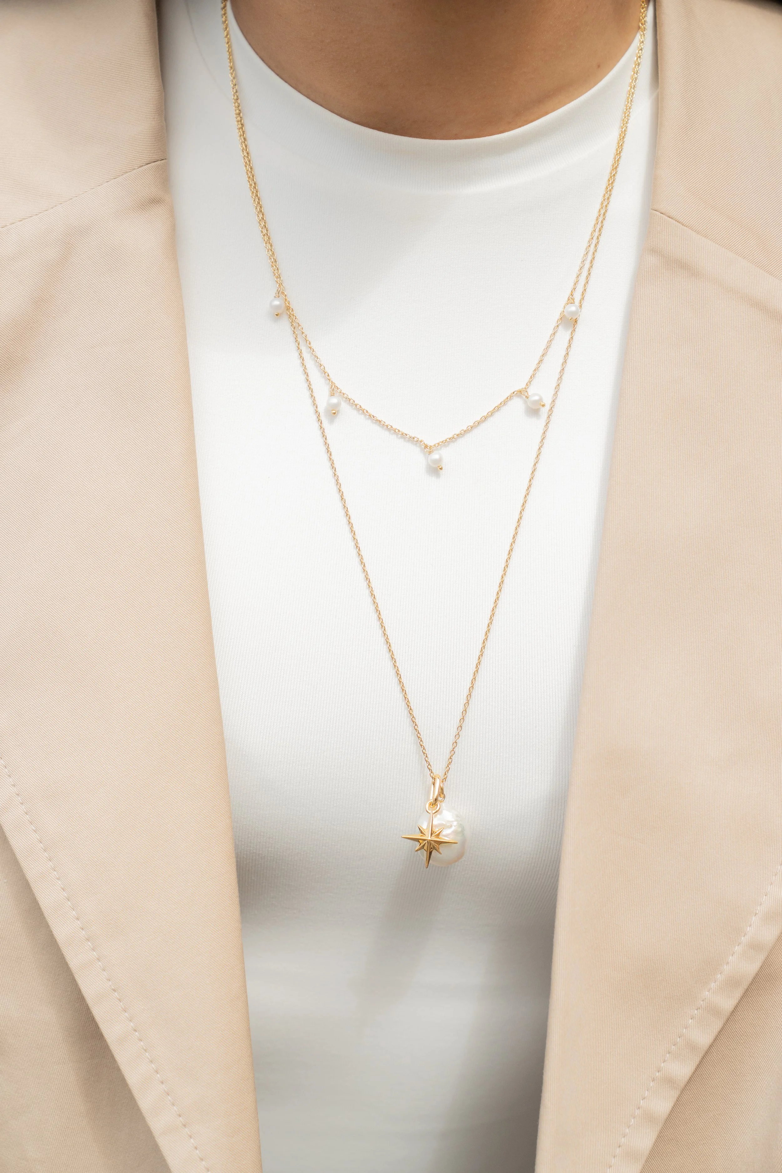 Close-up of a person wearing layered gold necklaces with pearls and a star-shaped pendant from Onna London, beige blazer, and white top.