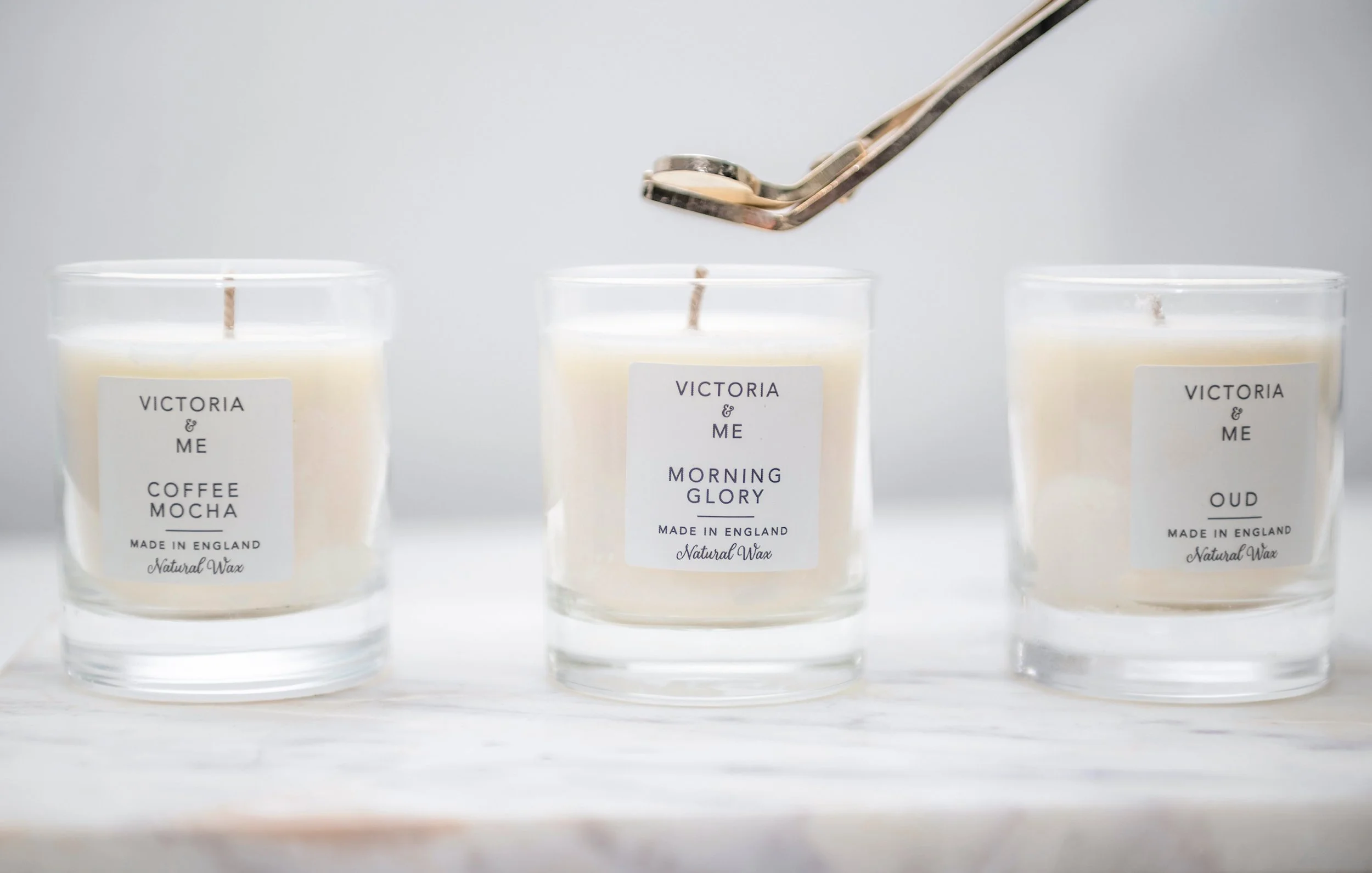 Three white candles in clear glass containers with labels reading Victoria & Me, placed on a white surface. One candle is being dipped into wax with a pair of tongs.