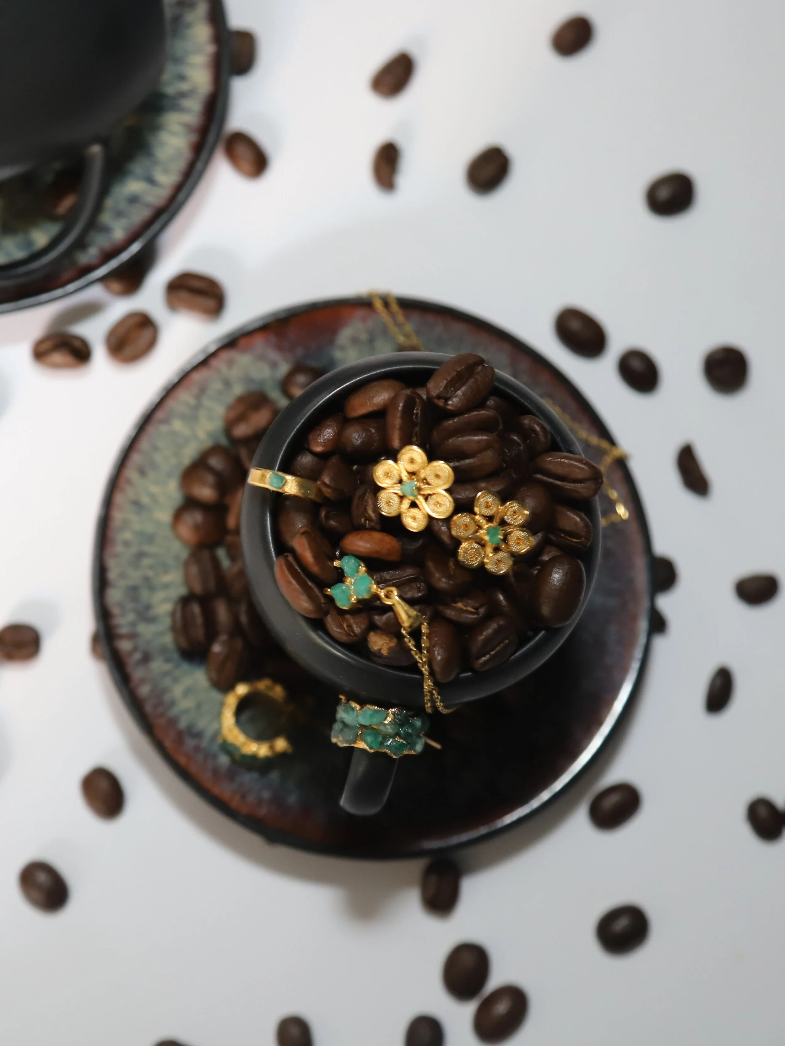 A black coffee mug filled with roasted coffee beans, placed on a decorative plate, with jewelry resting on the beans, surrounded by scattered coffee beans on a white background.