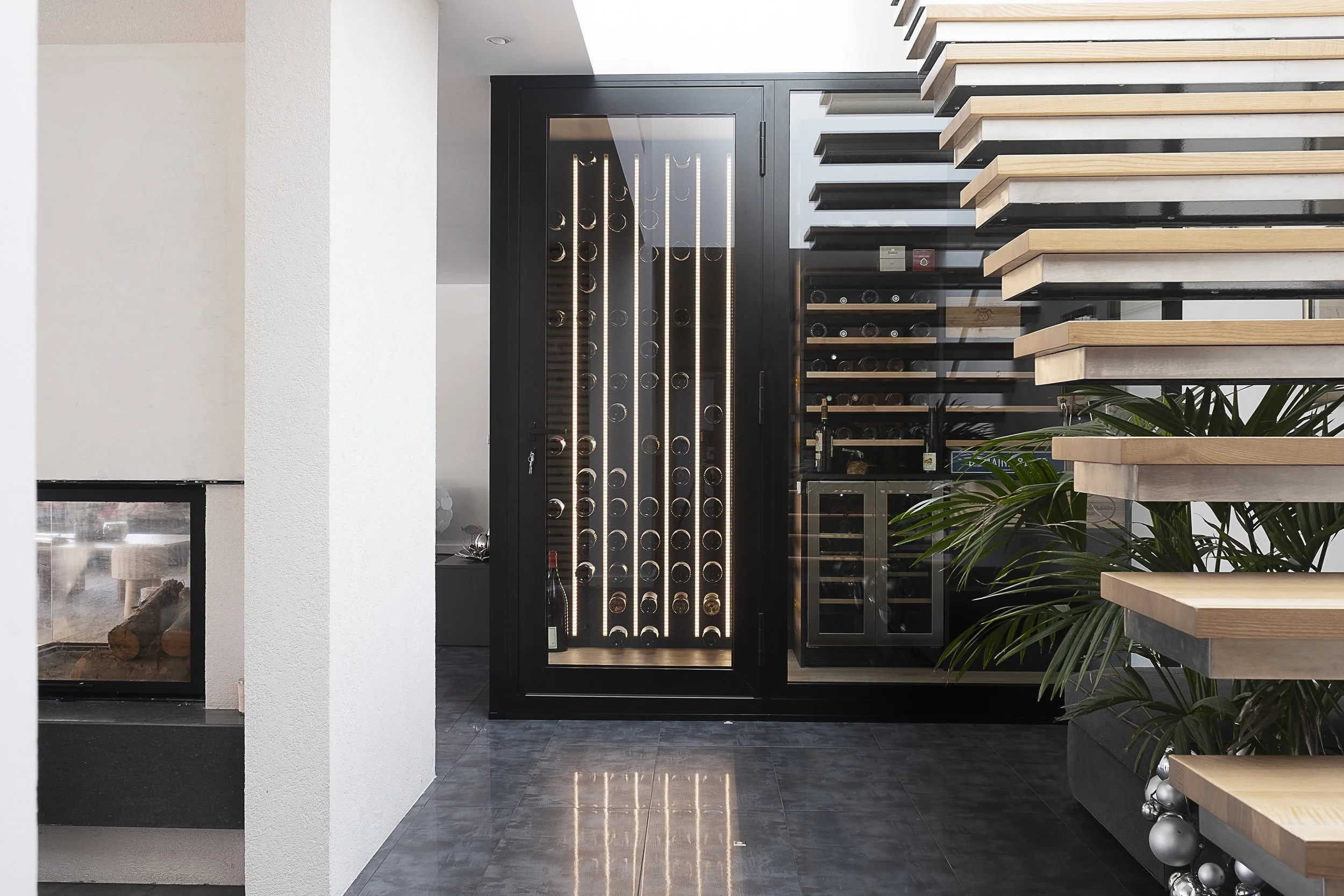 Cave à vin noire avec portes en verre, sapinée et éclairée intérieurement, située dans un intérieur moderne avec escaliers en bois et plantes vertes.