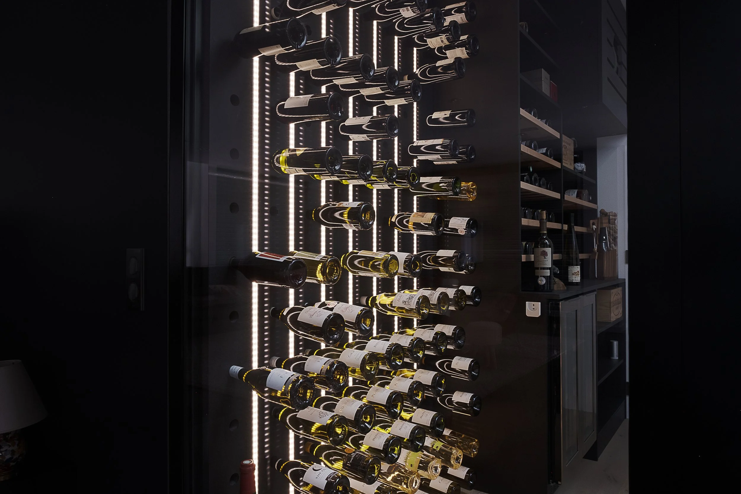 Une cave à vin moderne avec des bouteilles de vin alignées sur des rayonnages en miroir, éclairées par des lumières LED derrière un mur en verre noir.