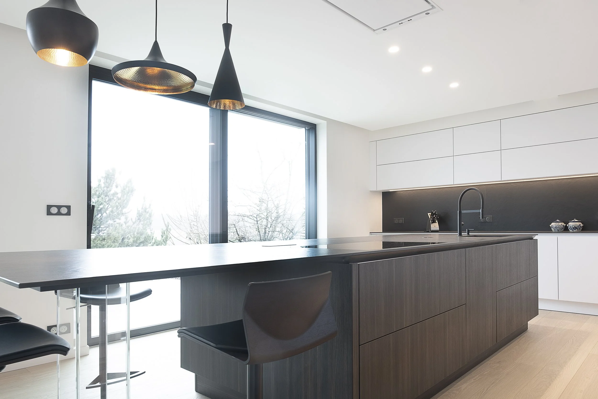 Cuisine moderne avec îlot en bois foncé, plan de travail noir, pôele intégré, évier avec robinet noir, lampe suspendue noire, grand fenêtre laissant entrer la lumière