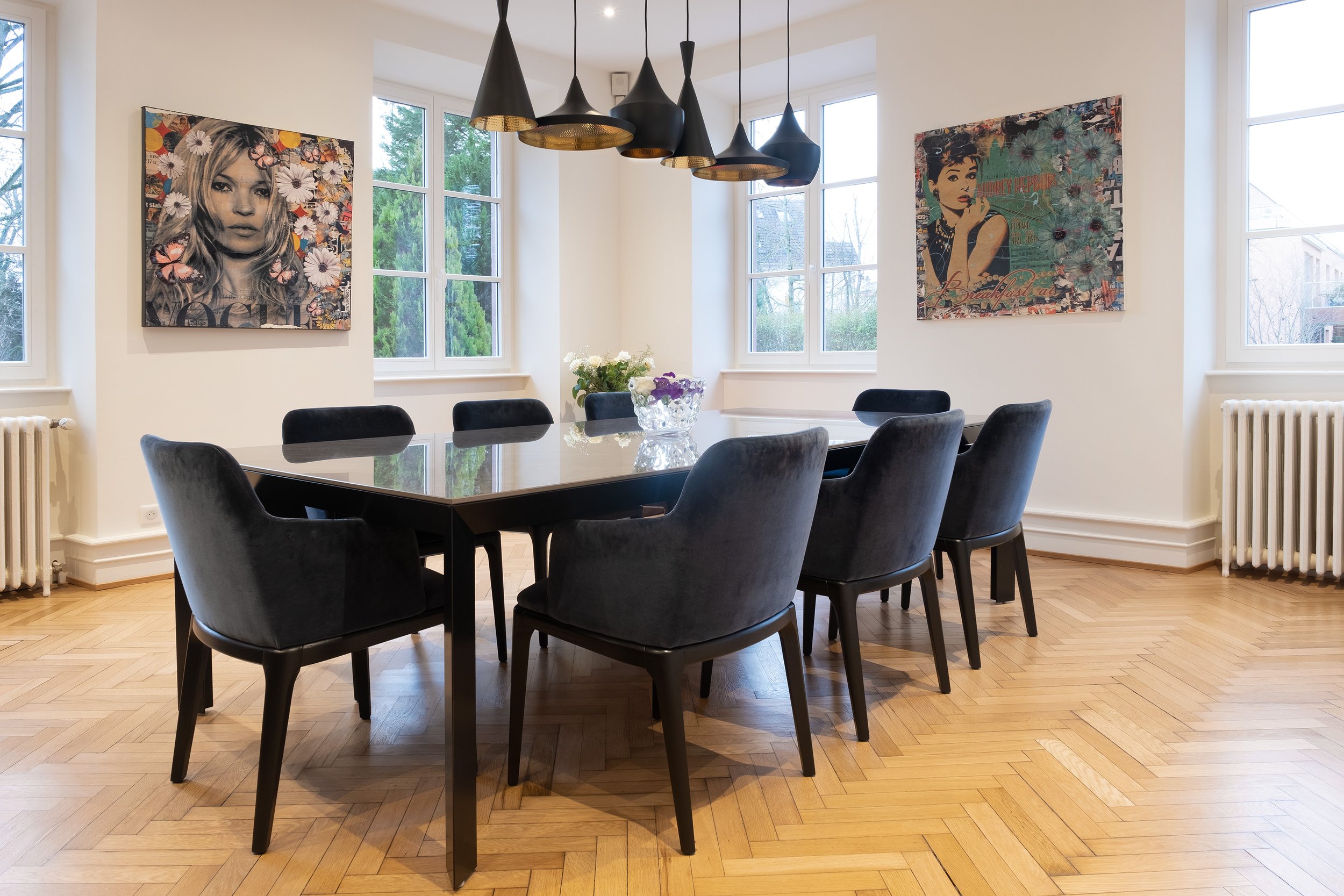 Salle à manger avec une grande table noire, six chaises en velours noir, deux paintings modernes accrochés au mur, trois fenêtres laissant entrer la lumière, des radiateurs et un lustre noir suspendu au plafond.