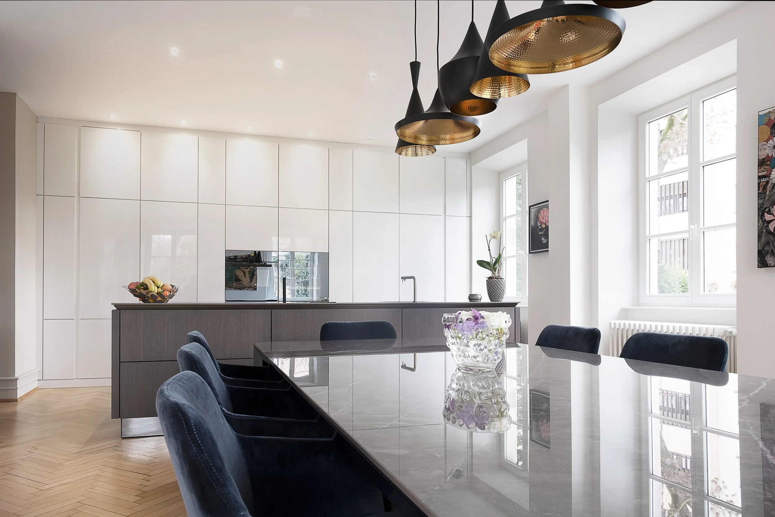 Salle à manger moderne avec un grand table en marbre, chaises noires, suspension noire et or, cuisine blanche avec îlot, grandes fenêtres laissant entrer la lumière, œuvres d'art sur les murs, décor épuré et lumineux.