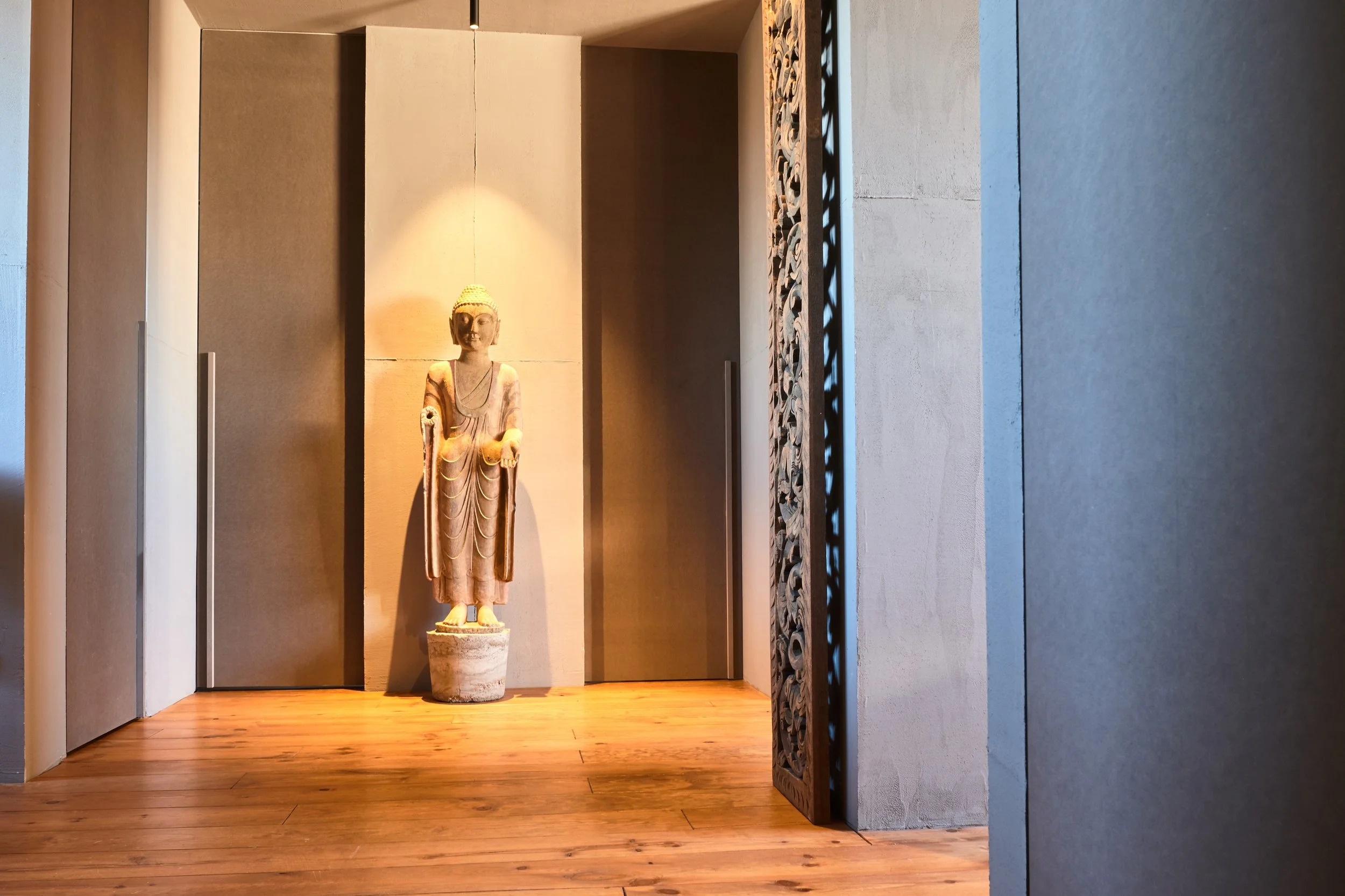 Statue de Bouddha dans une alcôve, éclairée, avec un sol en bois et un panneau en bois avec des motifs sculptés sur le côté.