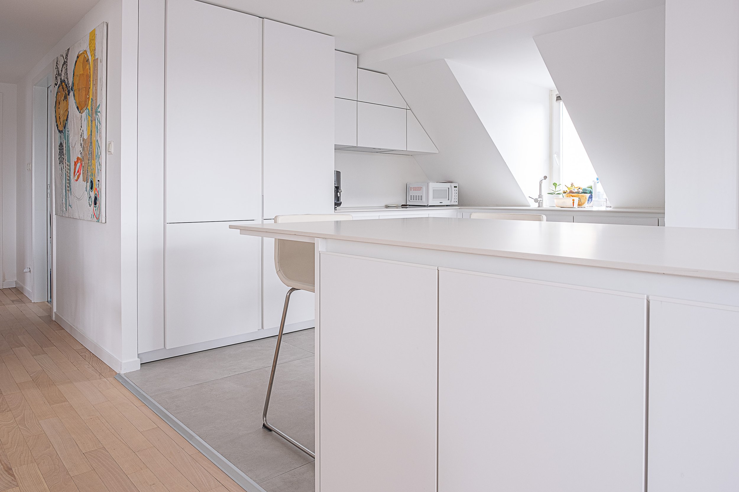 Cuisine moderne avec comptoir blanc, micro-ondes, évier, fenêtre avec lumière naturelle, murs et armoires blancs, tableau coloré sur le mur à gauche, sol en bois et carrelage gris.