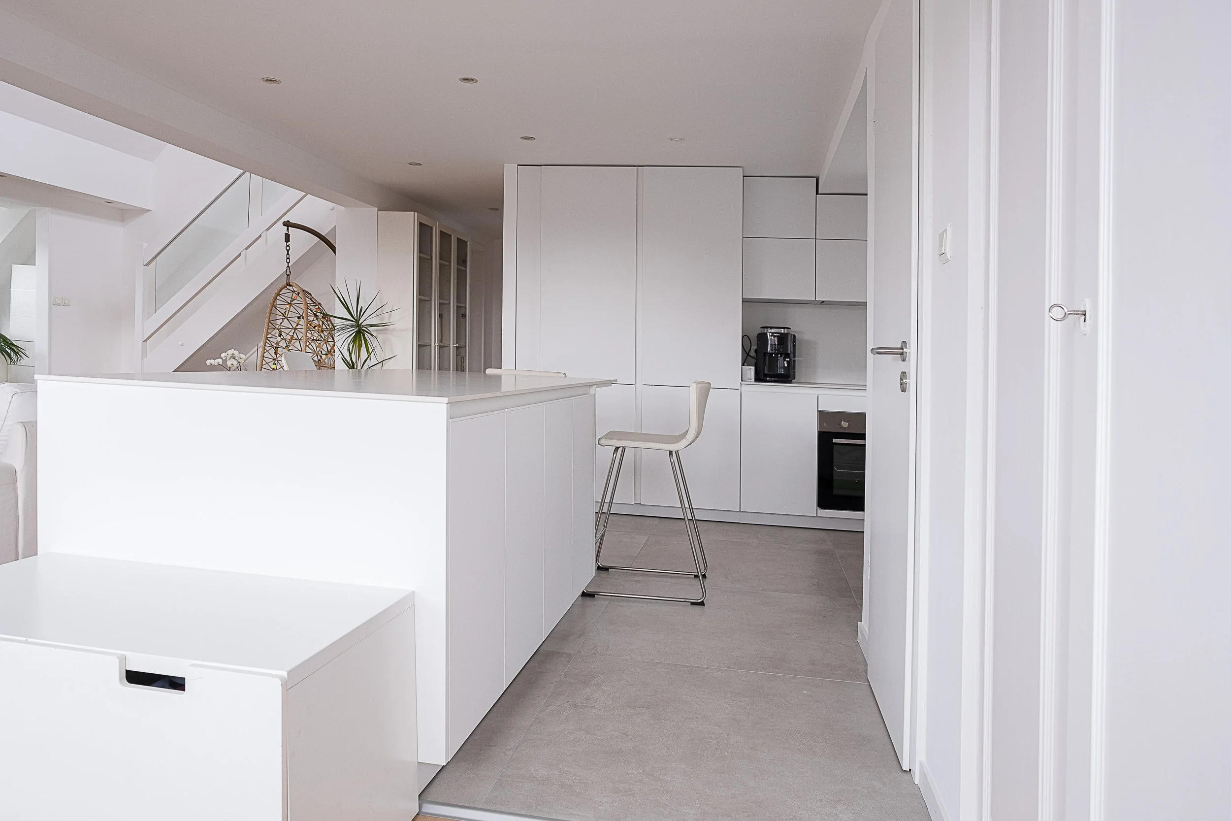 Cuisine entièrement blanche avec un îlot central, une chaise blanche avec structure métallique, un four intégré, une machine à café, et un décor moderne avec plantes vertes et lampe suspendue en osier.