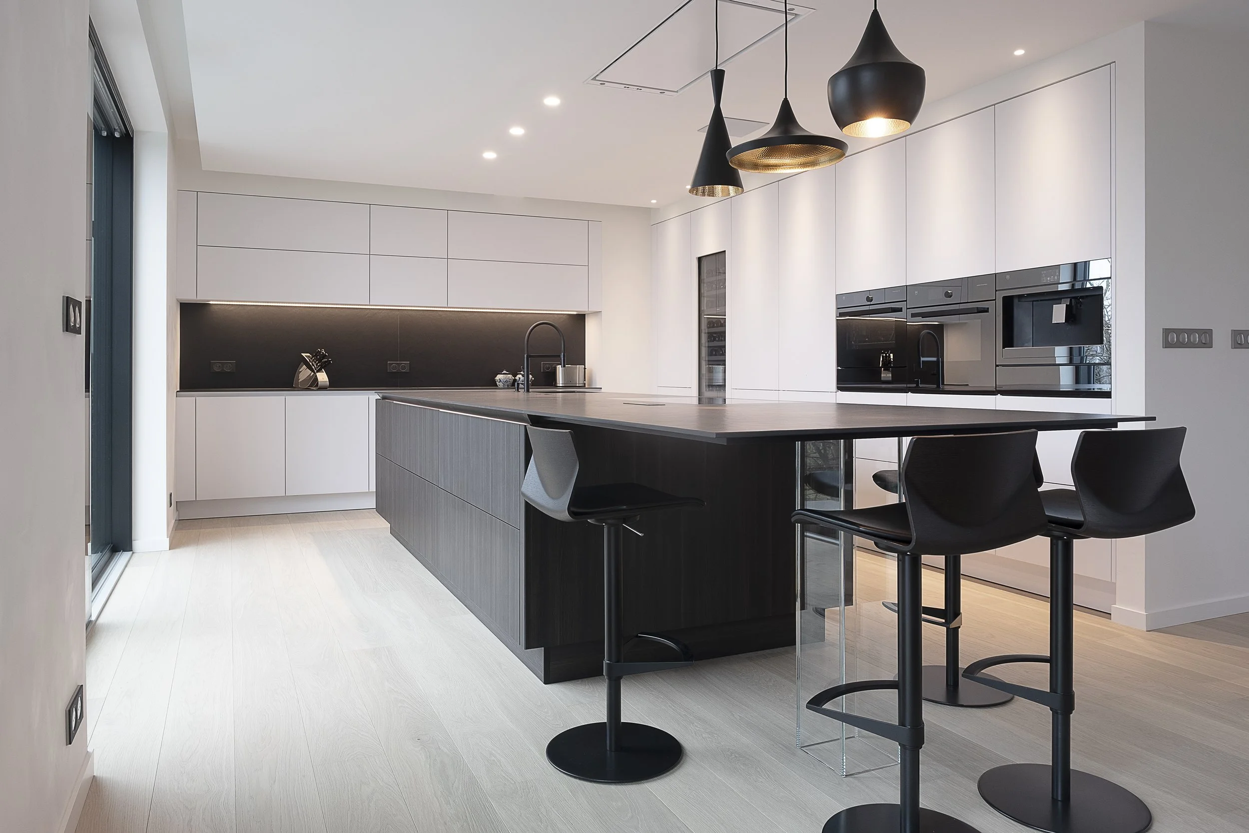 Cuisine moderne avec un îlot central en bois foncé, armoires blanches, plan de travail noir, suspendus noirs au plafond et chaises de bar noires.