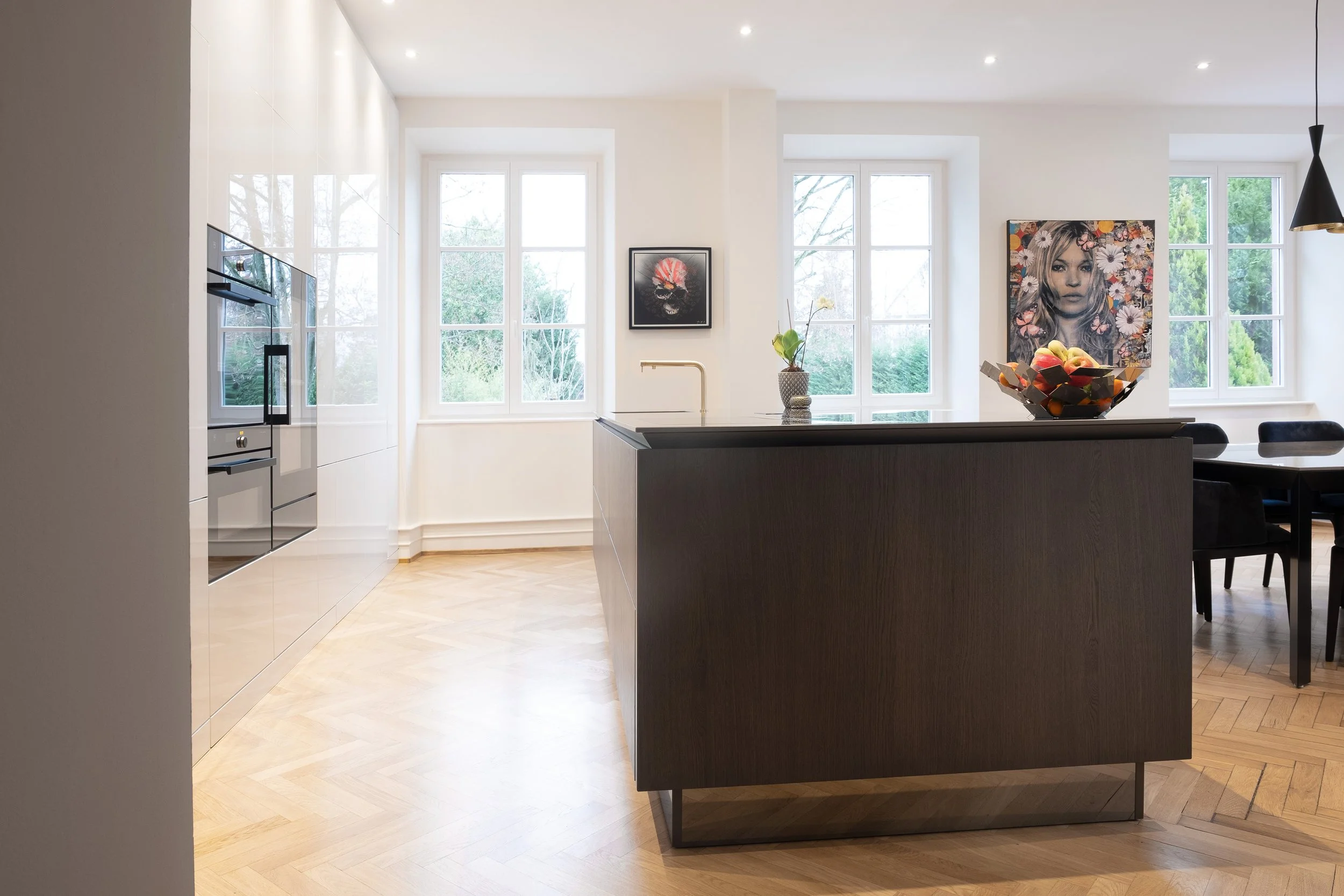 Cuisine moderne avec grand îlot en bois foncé, grandes fenêtres laissant entrer de la lumière naturelle, œuvres d'art sur les murs, et une table à manger en arrière-plan.