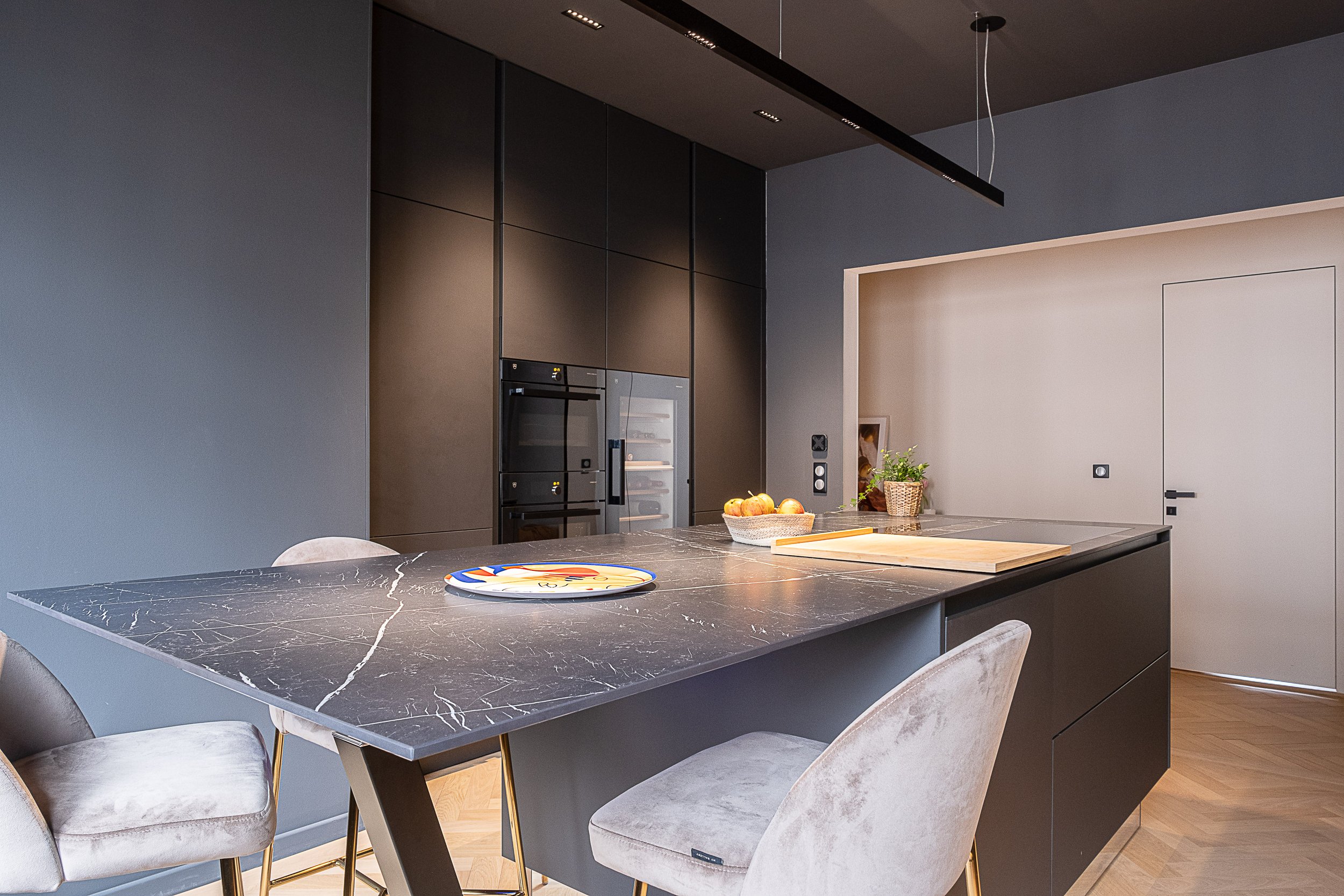 Cuisine moderne avec un grand plan de travail en marbre noir, des chaises en velours beige, et des appareils encastrés noirs et en verre, avec un décor minimaliste et épuré.