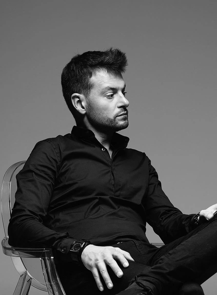 Un homme assis sur une chaise en plastique transparent, portant une chemise noire, regardant vers la droite, avec un fond gris