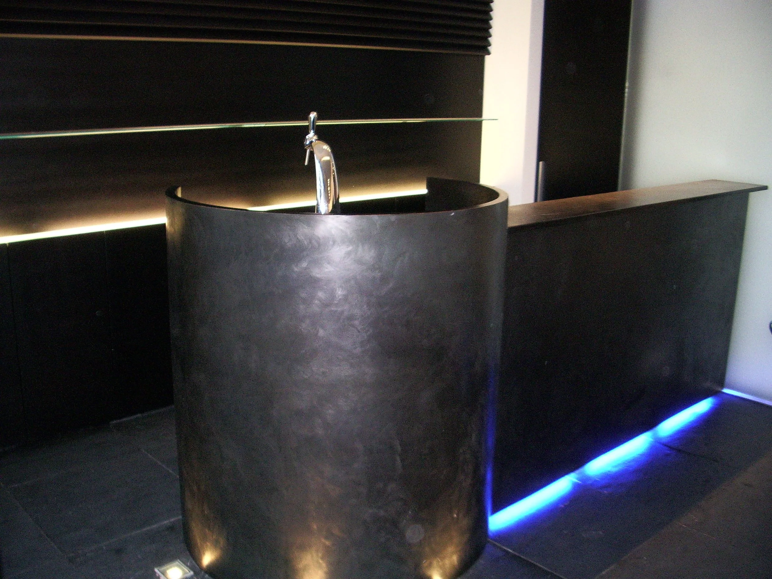 Une fontaine d'eau noire avec éclairage bleu en dessous, située dans une pièce moderne avec des murs sombres et un sol en carrelage noir.