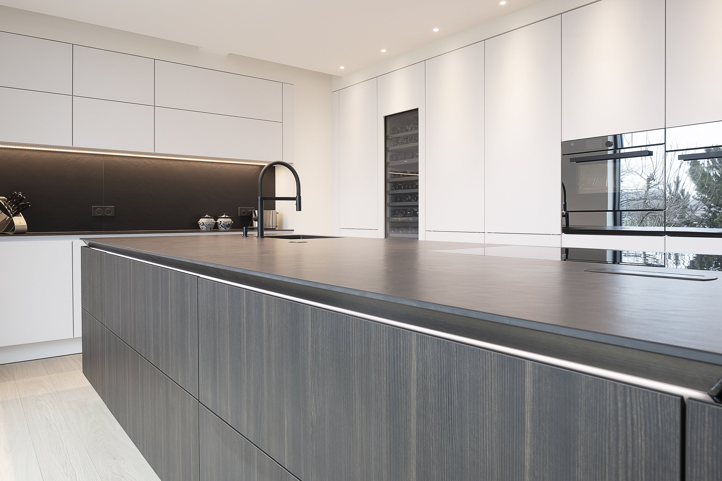 Cuisine moderne avec un grand îlot central en bois foncé, façades en bois clair, murs blancs, un réfrigérateur encastré, et une fenêtre avec vue sur des arbres.