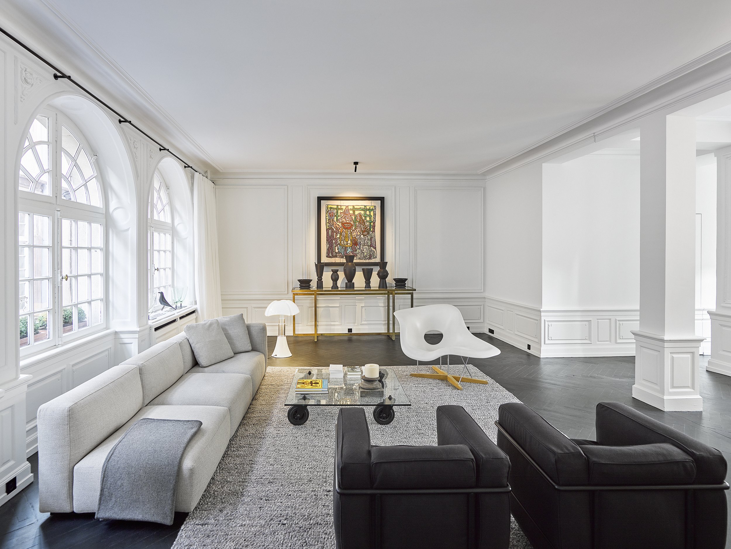 Salon moderne avec canapé gris, fauteuil blanc, canapé noir, table en verre, lampadaire blanc, œuvres d'art, grande fenêtre, décor blanc, sol en bois foncé.