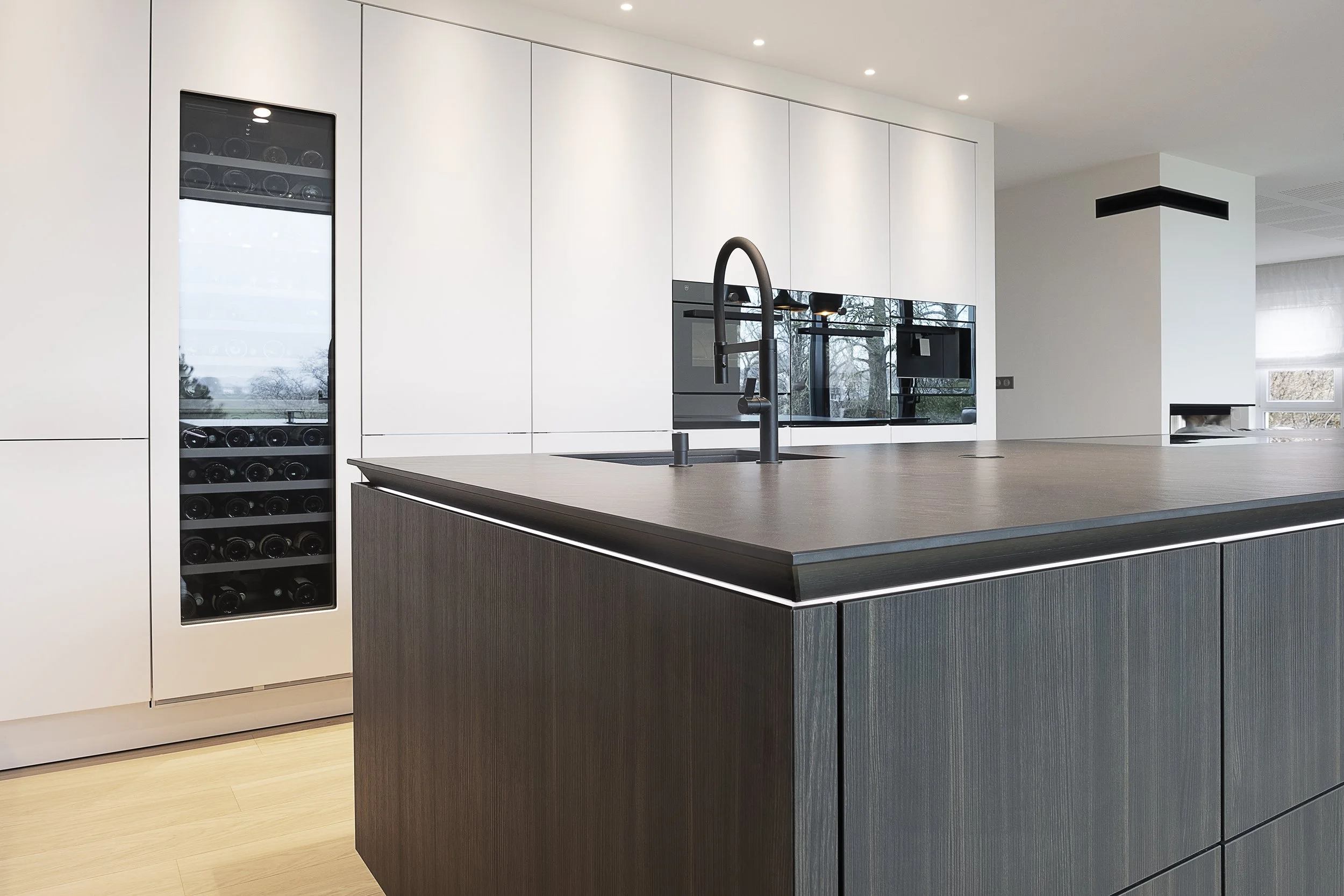 Cuisine moderne avec un îlot central en bois foncé, évier et robinet noir, murs blancs, un réfrigérateur encastré en verre noir et des fenêtres laissant entrer la lumière naturelle.