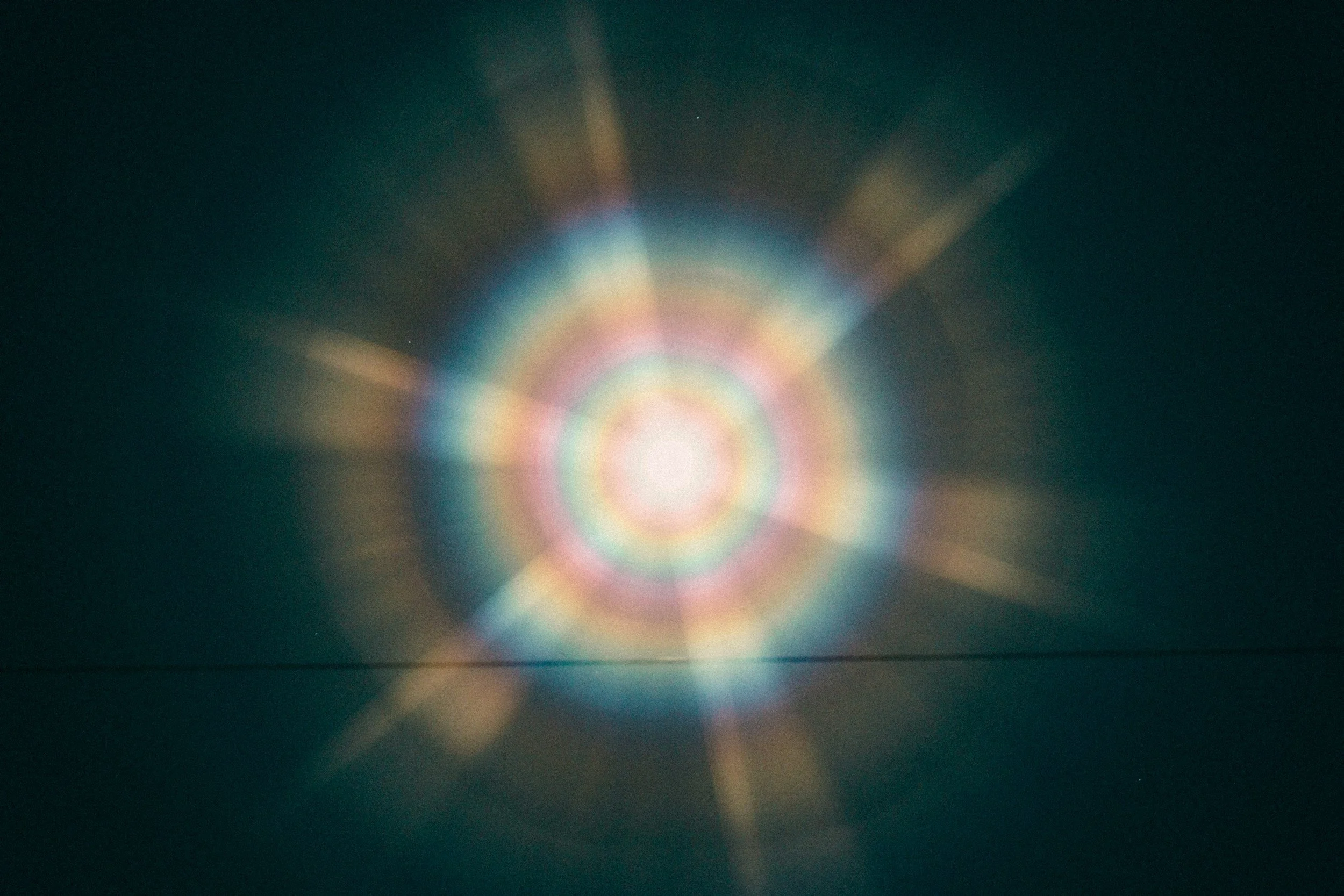 Colorful rainbow light rays radiate outward in a circular pattern against a dark background.