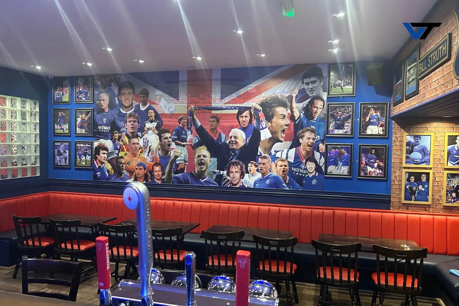 Interior of a sports bar decorated with framed photos and a large mural of soccer players, including a central figure holding a scarf, with tables and chairs in the foreground.