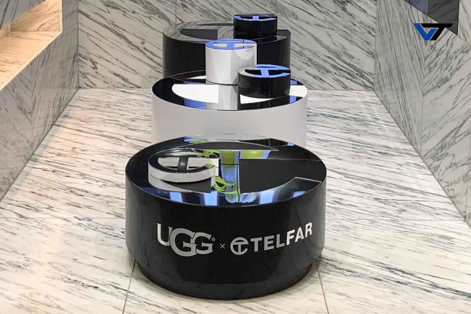 Metal sculpture of layered circular shapes with open spaces, featuring logos for UGG and TelFar, placed on a marble floor with marble walls.