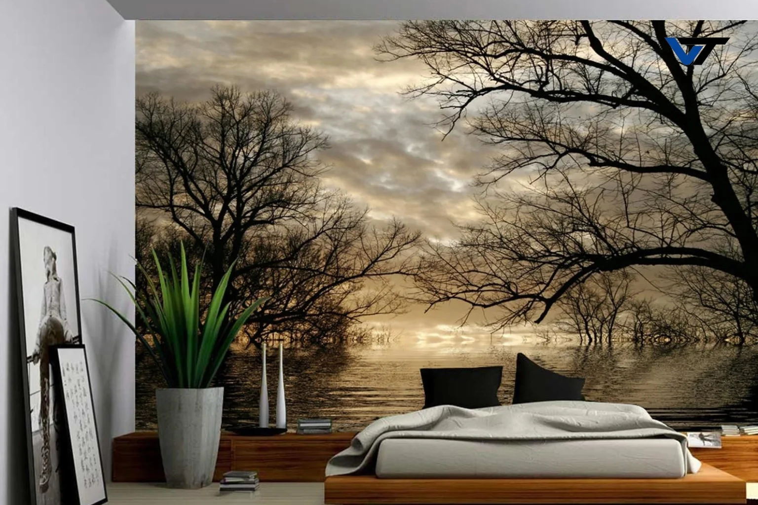 Interior of a room with a large nature-themed mural of leafless trees over a body of water at sunset, two black chairs, a white sofa with a blanket, a large potted plant, and framed artwork leaning against the wall.
