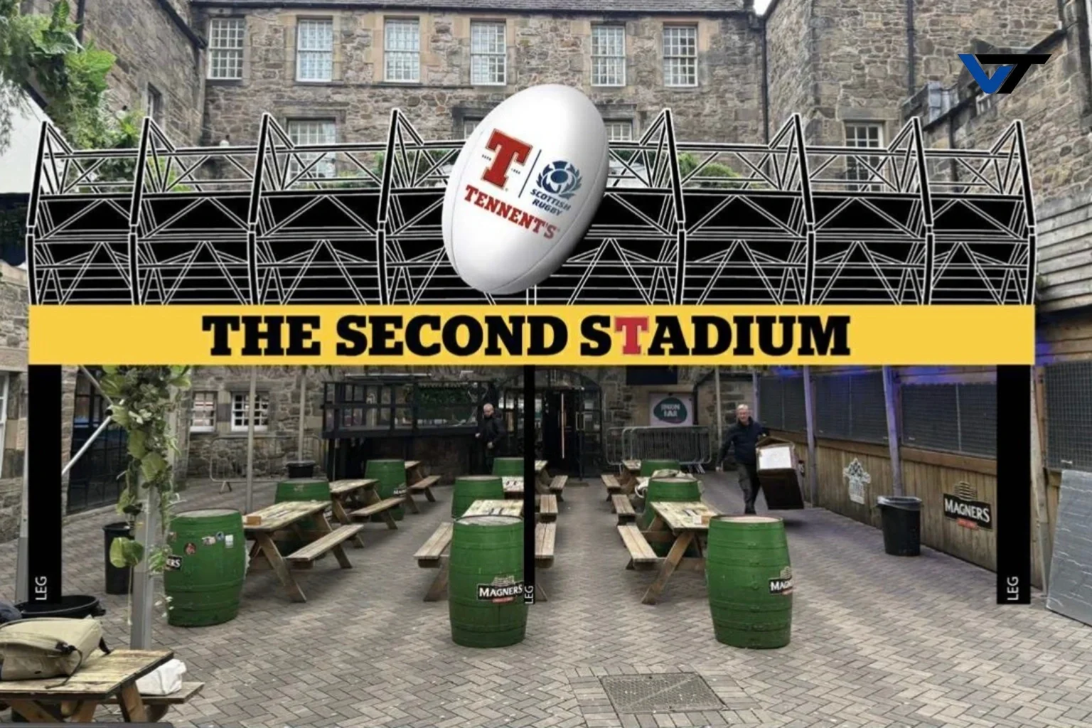 A stadium entrance with a large sign reading 'The Second Stadium' featuring a rugby ball with sponsorship logos. There are green barrels and wooden picnic tables in front, along with a few people walking.