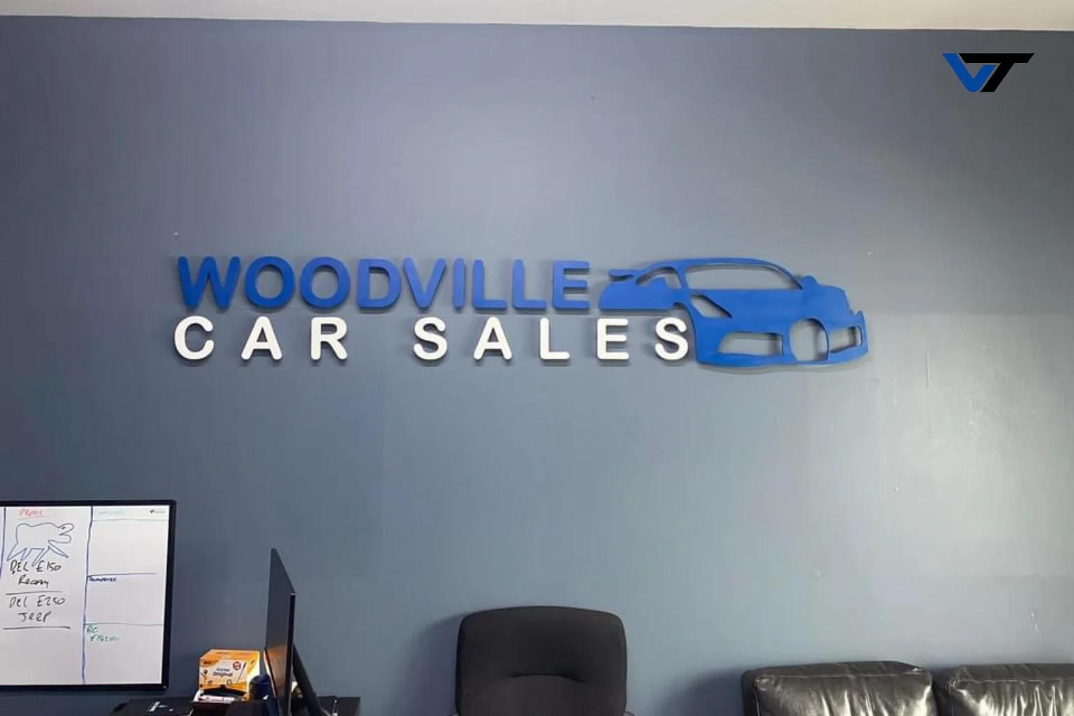 Photo of a wall sign that reads 'Woodville Car Sales' with a stylized blue car outline on the right side. The sign is in blue and white letters, with a small logo in the top right corner.