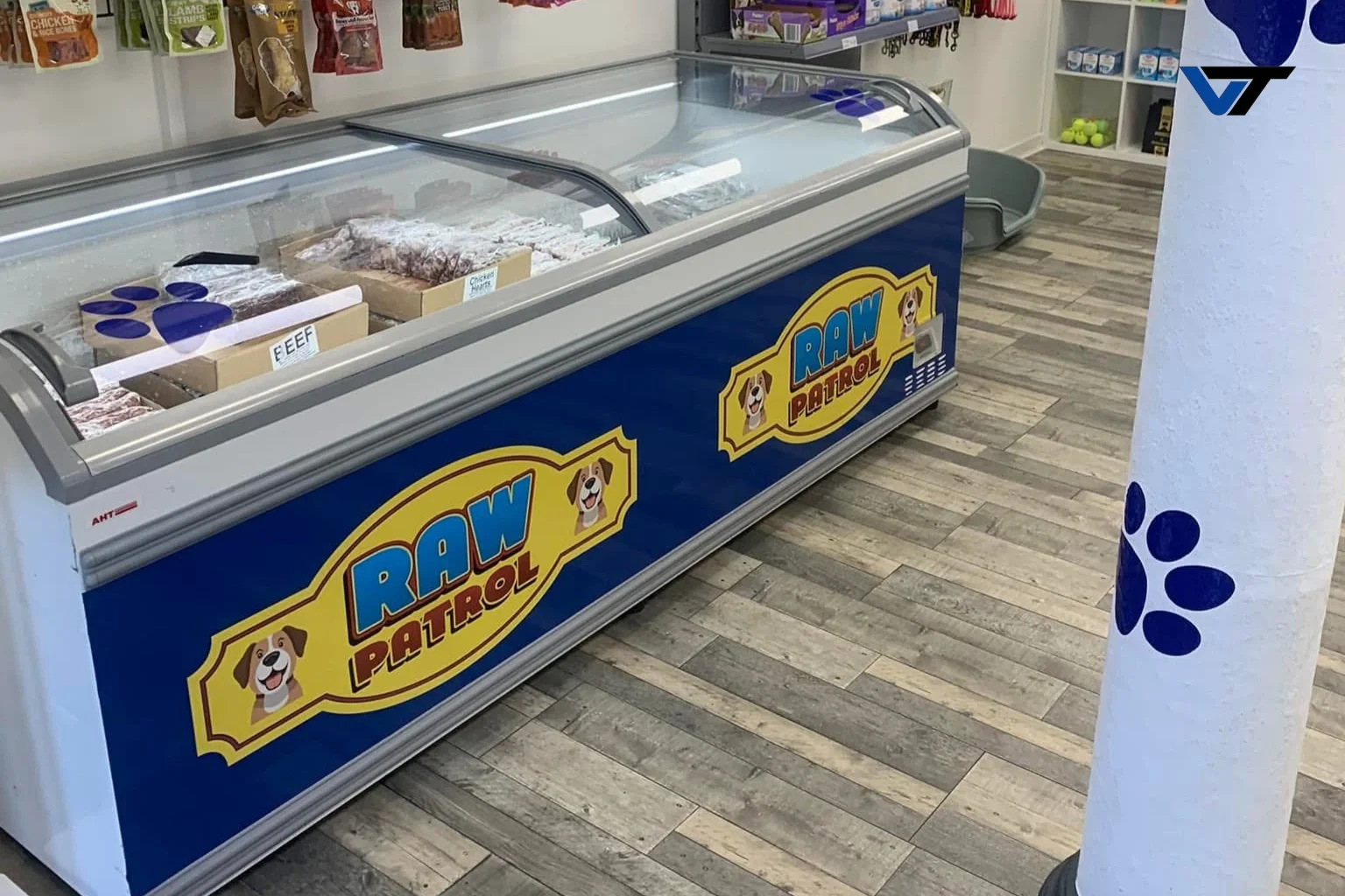 Ice cream freezer with a "Raw Patrol" logo and a dog illustration, in a pet store or zoo store, on a wood floor with shelves of pet products in the background.