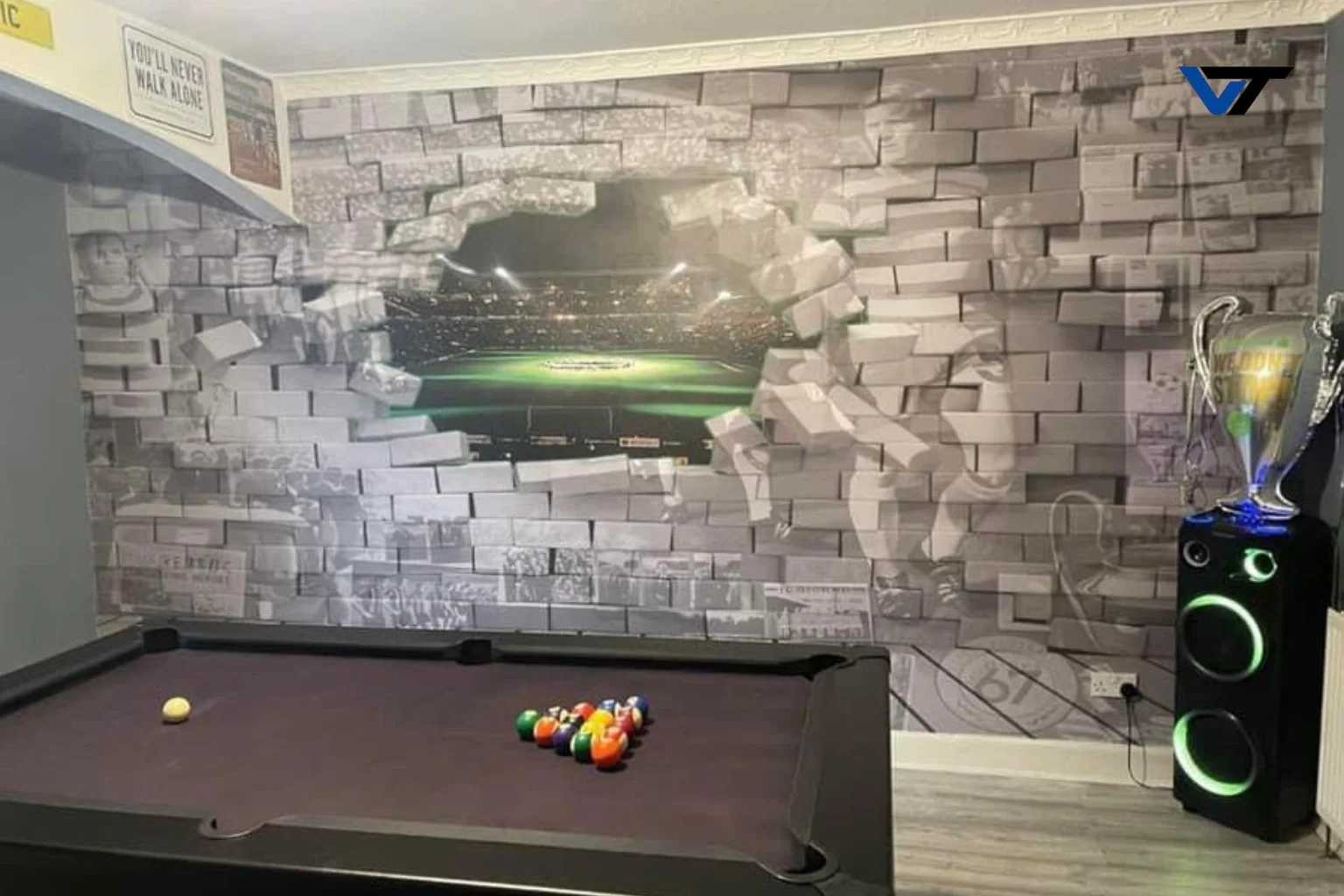 A pool table with billiard balls racked at one end, a cue ball, and a mural of a stadium with a hole in the brick wall creating a 3D effect. There are signs and a large speaker nearby.