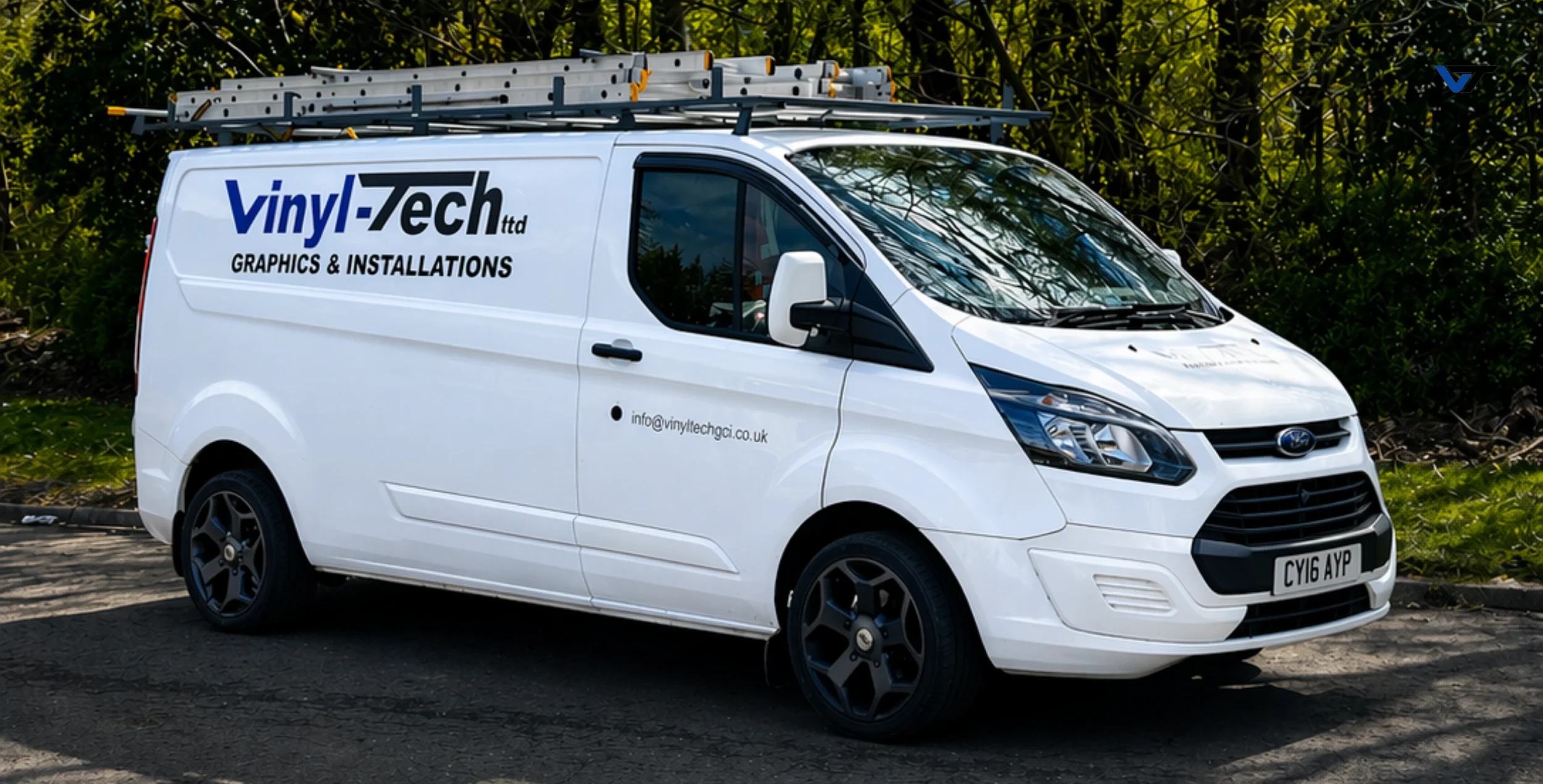 White van with vinyl-tek graphics, ladder on roof, parked outdoors near greenery.