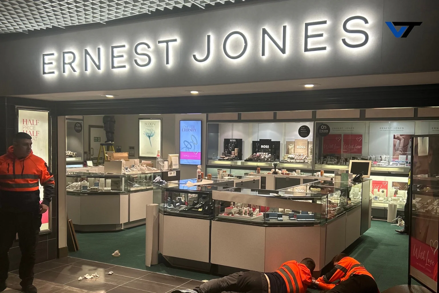Inside a jewelry store named Ernest Jones, with displays of jewelry, two workers in orange and black uniforms attending to a situation on the floor, and a security guard standing near the entrance.