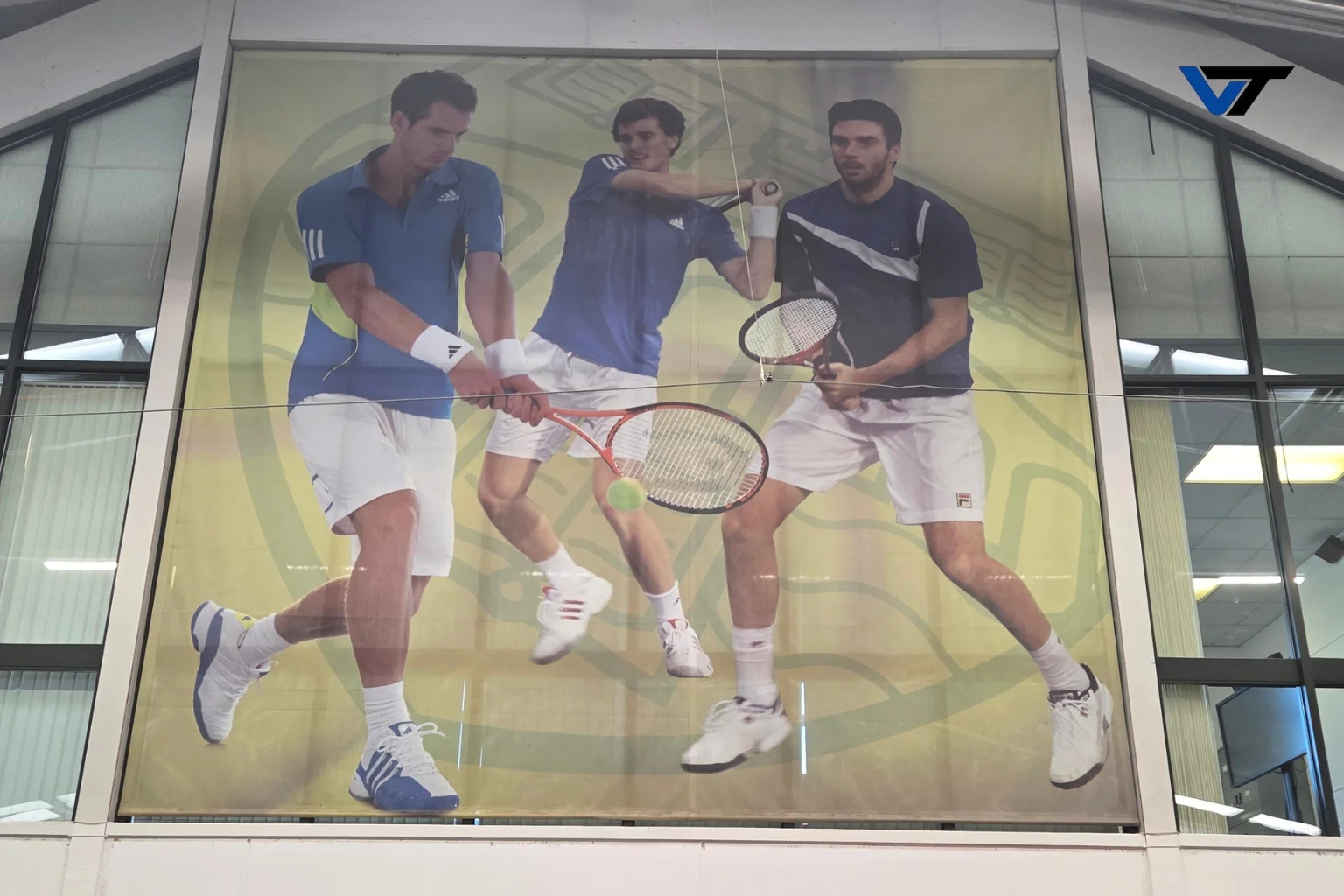 Large poster of three male tennis players in action, wearing blue and white sports attire, holding tennis rackets, on a yellow abstract background, displayed in a modern indoor setting.